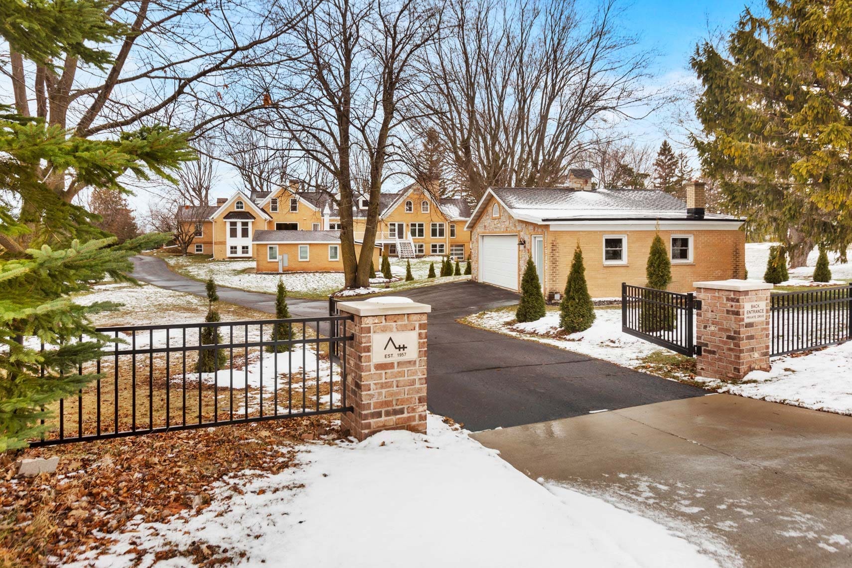 Residential real estate property in winter showcasing a tan single-family home with a single-car garage and paved driveway. The property is surrounded by trees, features a metal gate, and is accented by brick pillars at the entrance.