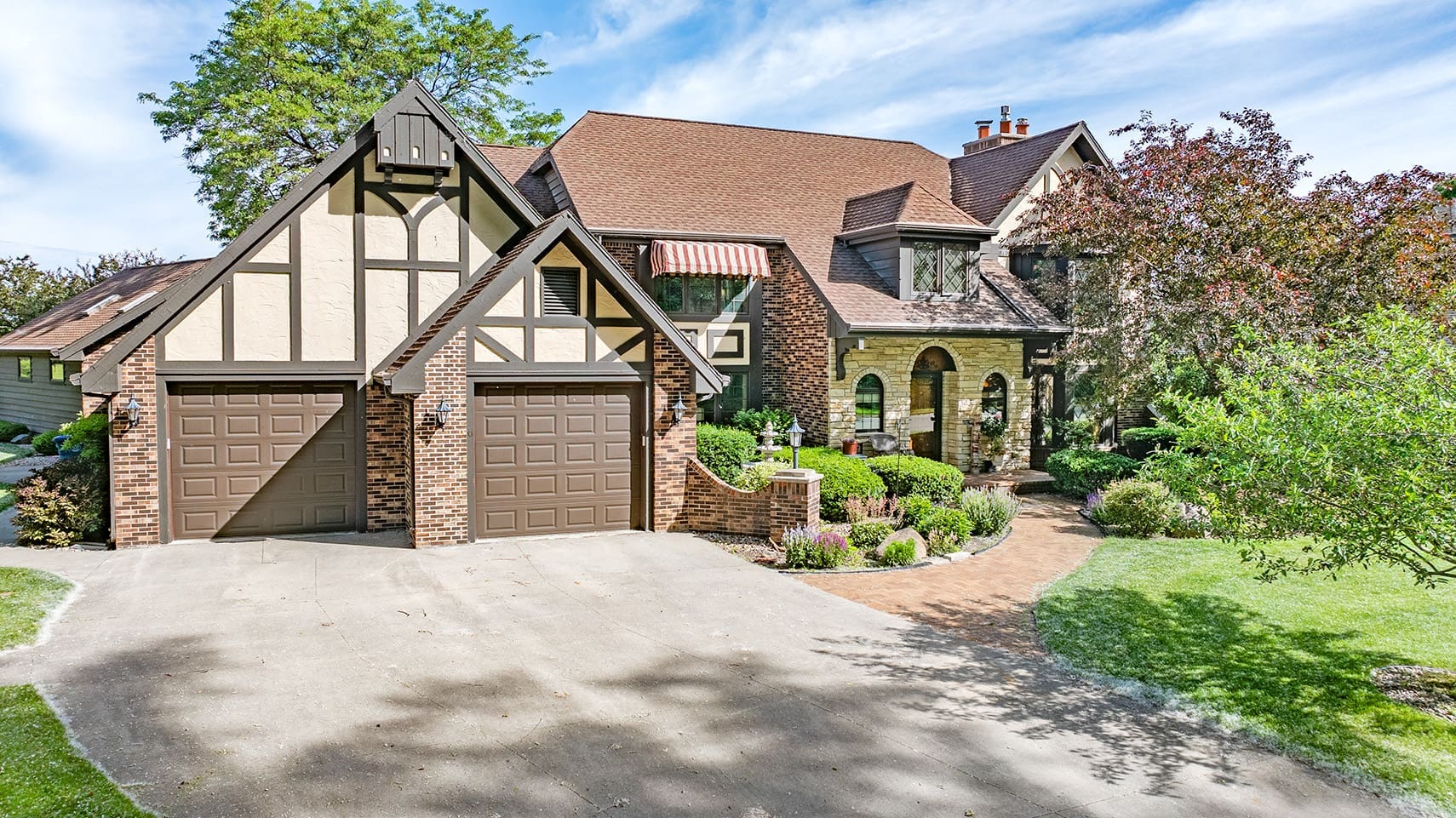 Tudor-style residential property showcasing brick and wood facade with two brown garage doors, manicured garden, and a curved walkway leading to the entrance for real estate listing.