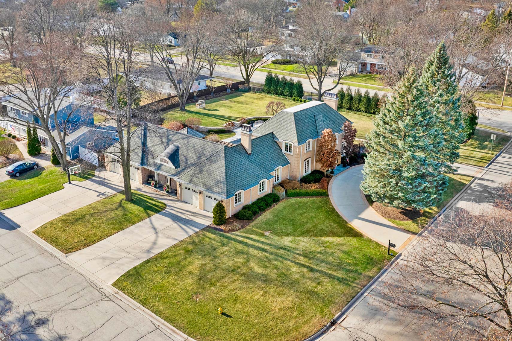 Aerial photo of a suburban residential property featuring a two-story house with a green roof, expansive driveway, lush lawn, and mature trees, highlighting its prime corner location in a neighborhood setting.