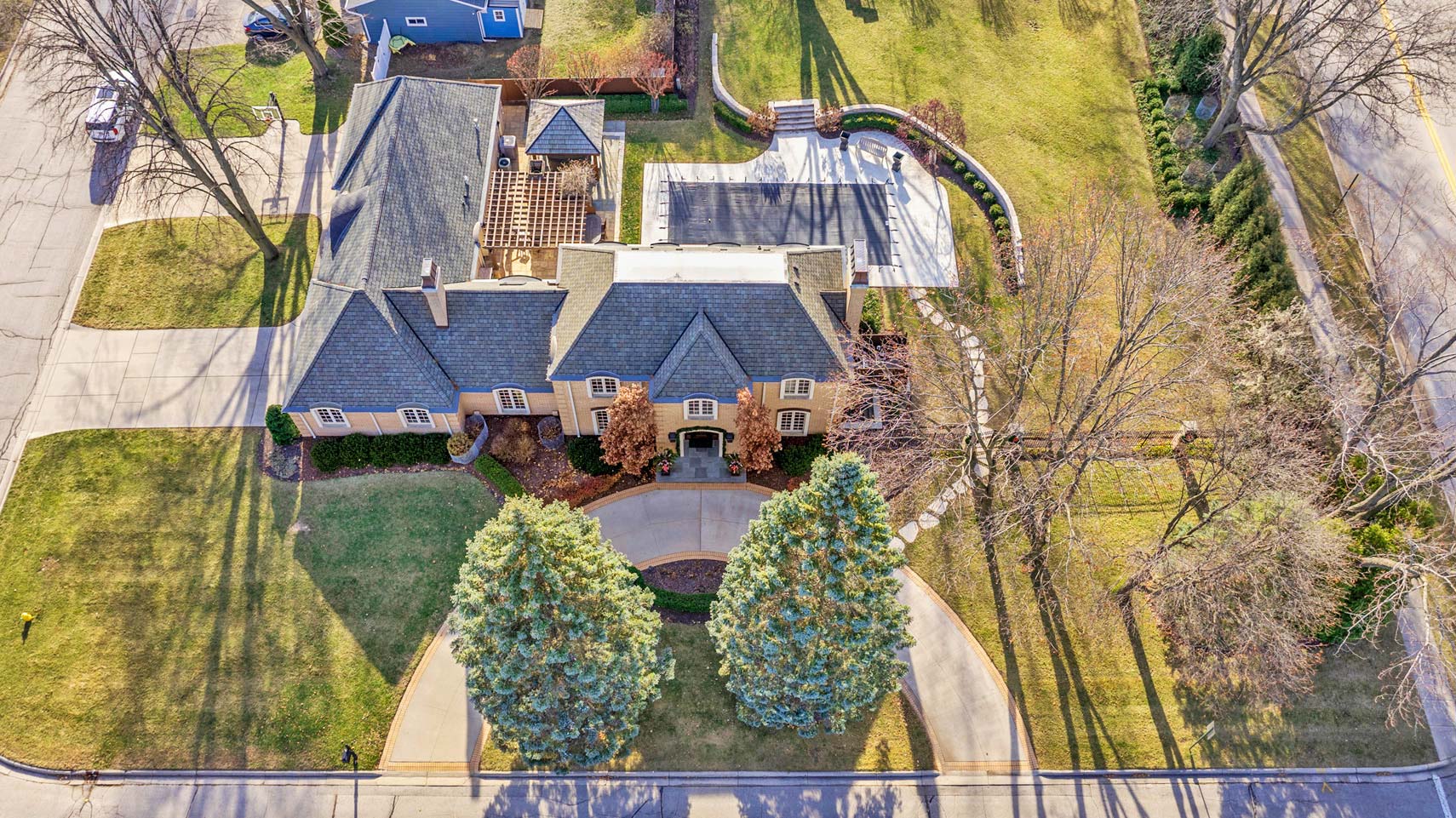 Aerial real estate photo of a luxury residential property with circular driveway, showcasing expansive green lawns and mature trees, at a road intersection.