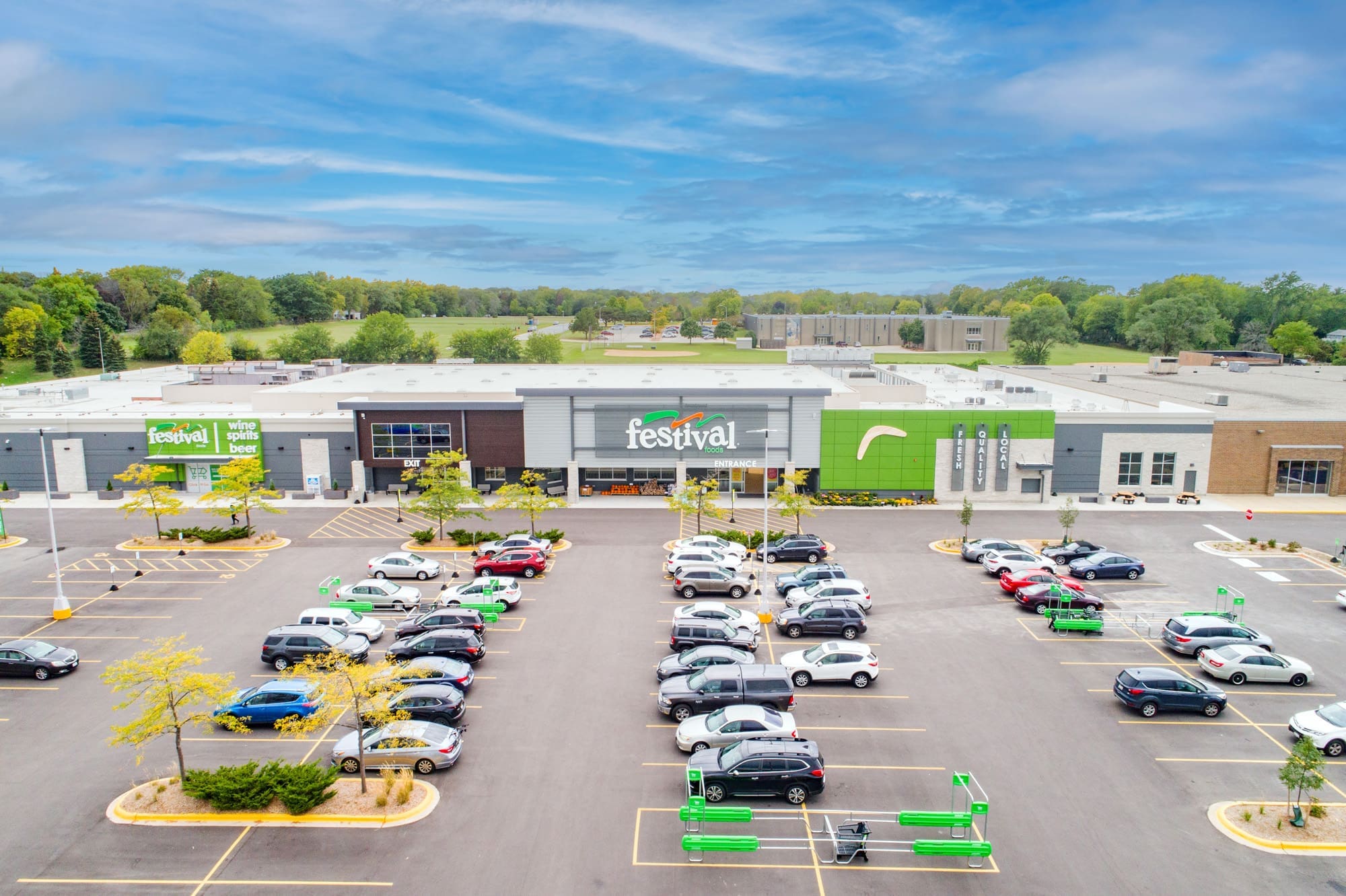 Aerial commercial real estate photo of Festival Foods grocery store, parking lot with vehicles, and landscaped trees under a partly cloudy sky. Aerial commercial real estate photo of Festival Foods grocery store, parking lot with vehicles, and landscaped trees under a partly cloudy sky.