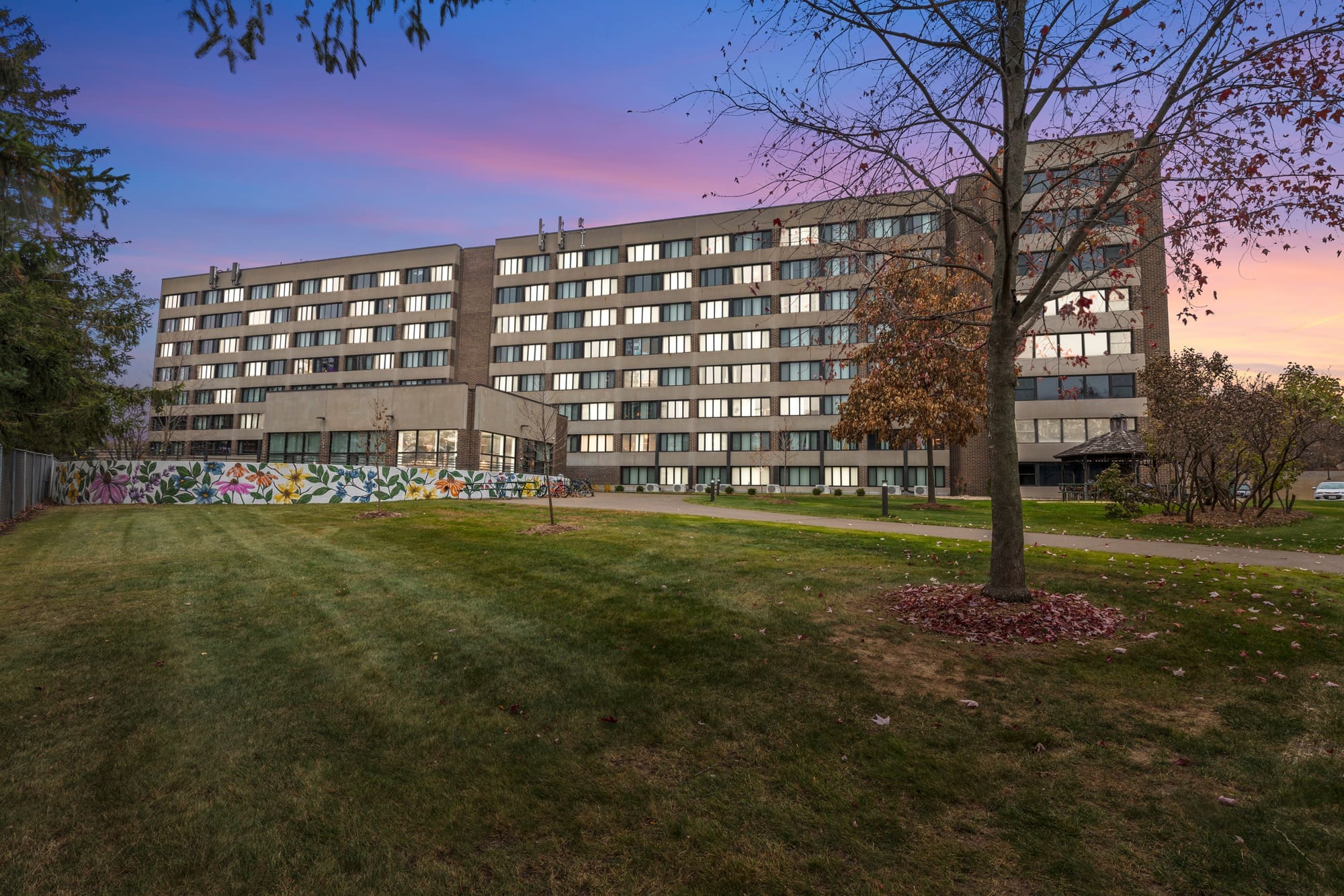 Modern multi-story residential apartment building with mural, landscaped lawn, and sunset lighting—ideal real estate photography. Modern multi-story residential apartment building with mural, landscaped lawn, and sunset lighting—ideal real estate photography.