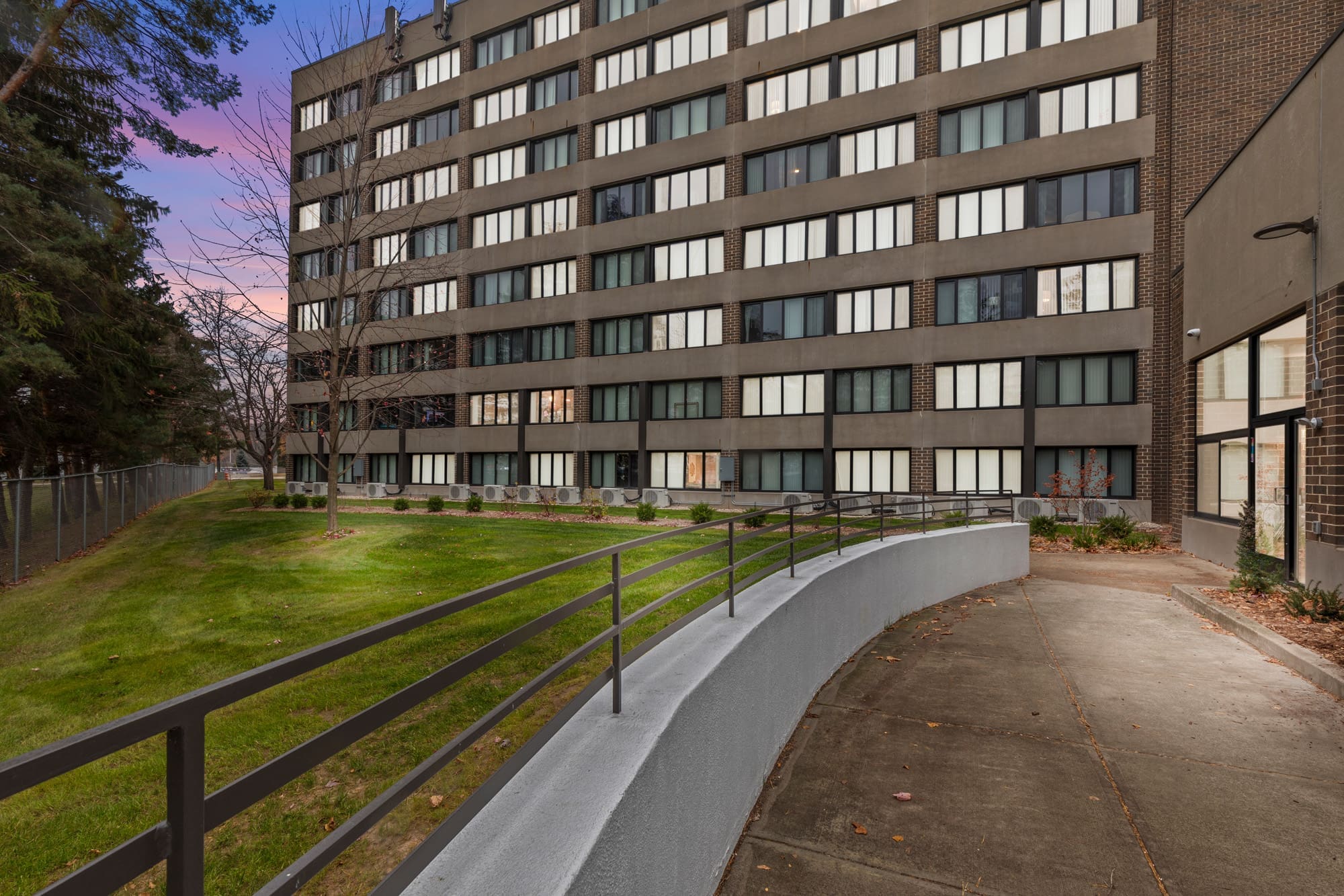 Residential apartment complex featuring multiple stories and large windows, landscaped lawn, walkway, and mature trees at dusk.