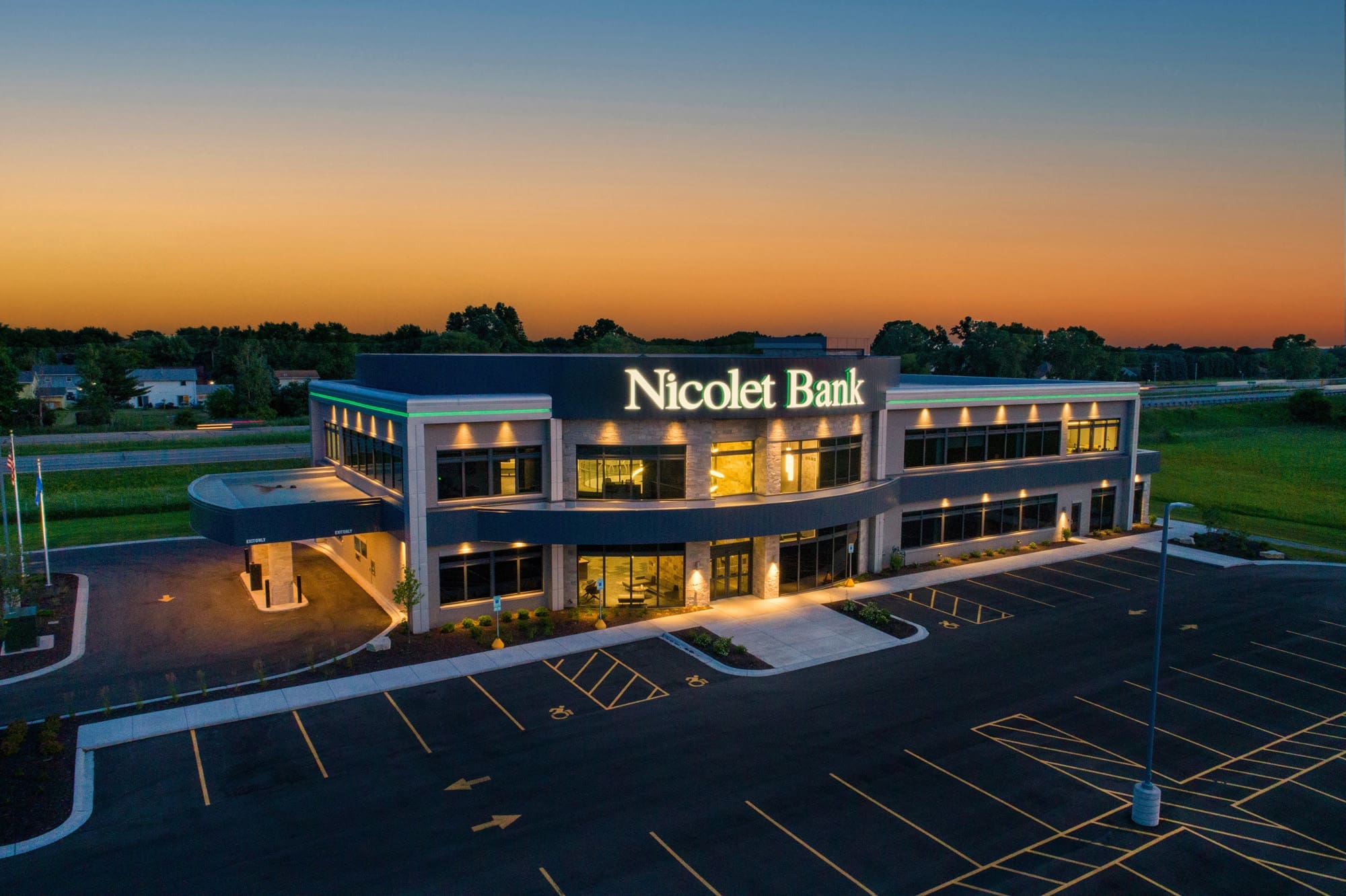 Commercial real estate photo of a modern Nicolet Bank building with large windows, lit at dusk, empty parking lot, sunset backdrop.