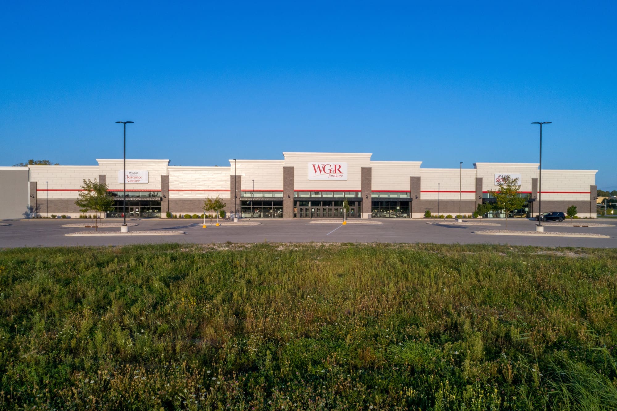 Commercial property for sale: vacant retail store with WGR signage, expansive parking area, overgrown landscaping, clear sky.