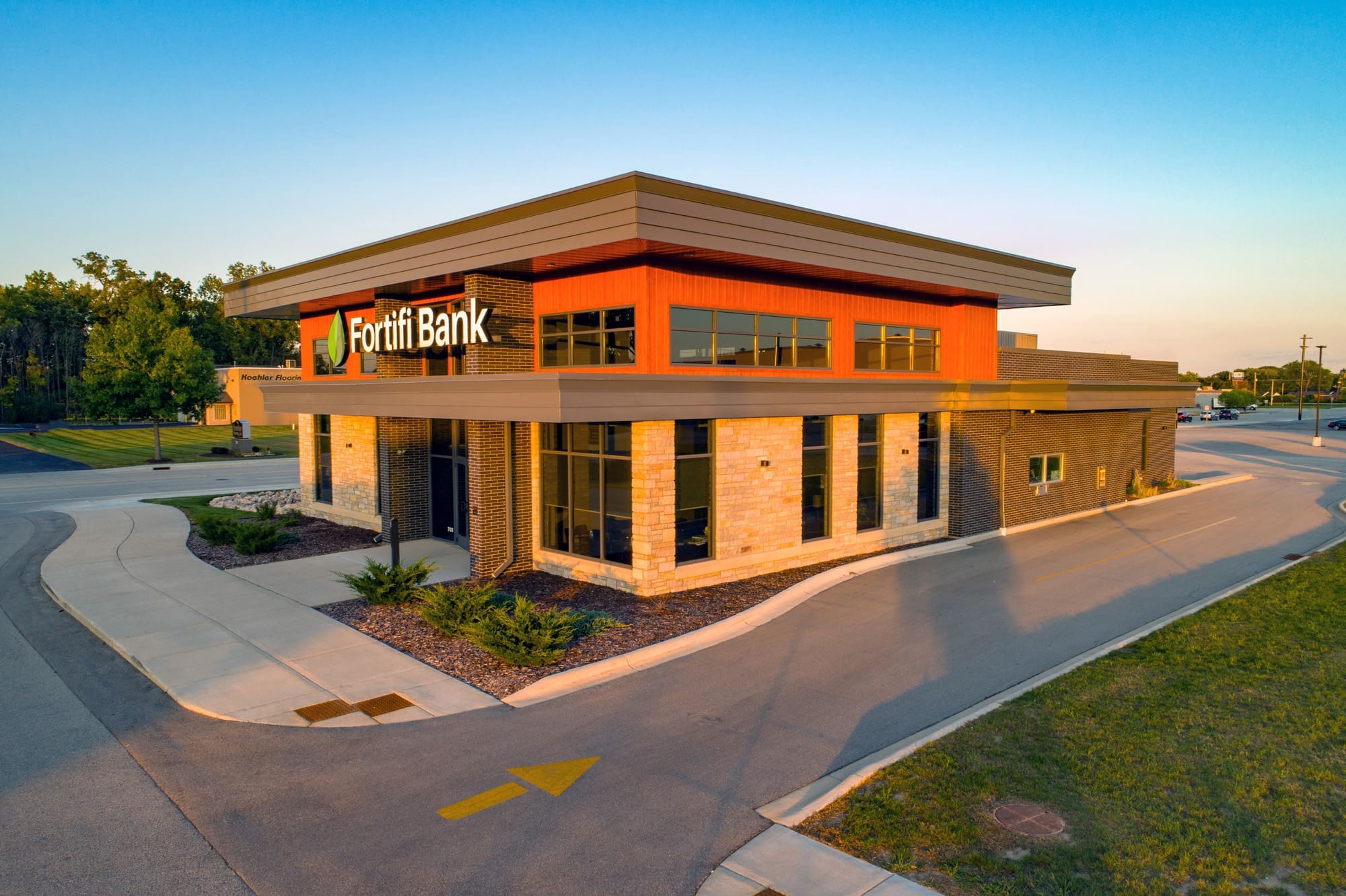 Commercial real estate photo of a modern, single-story Fortifi Bank with large windows, red and beige facade, and parking lot at sunset.