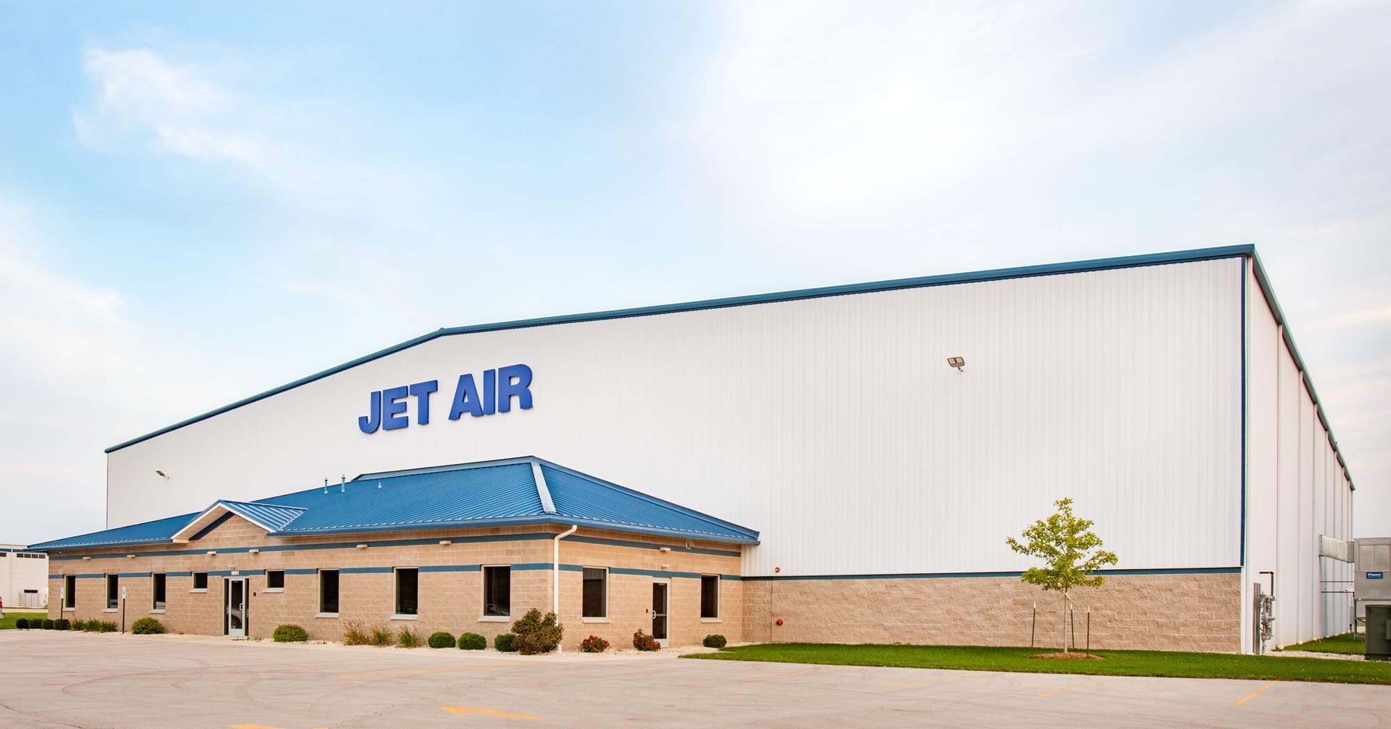 Commercial property for sale featuring a large white industrial building with blue accents, JET AIR signage, paved lot, and landscaping.