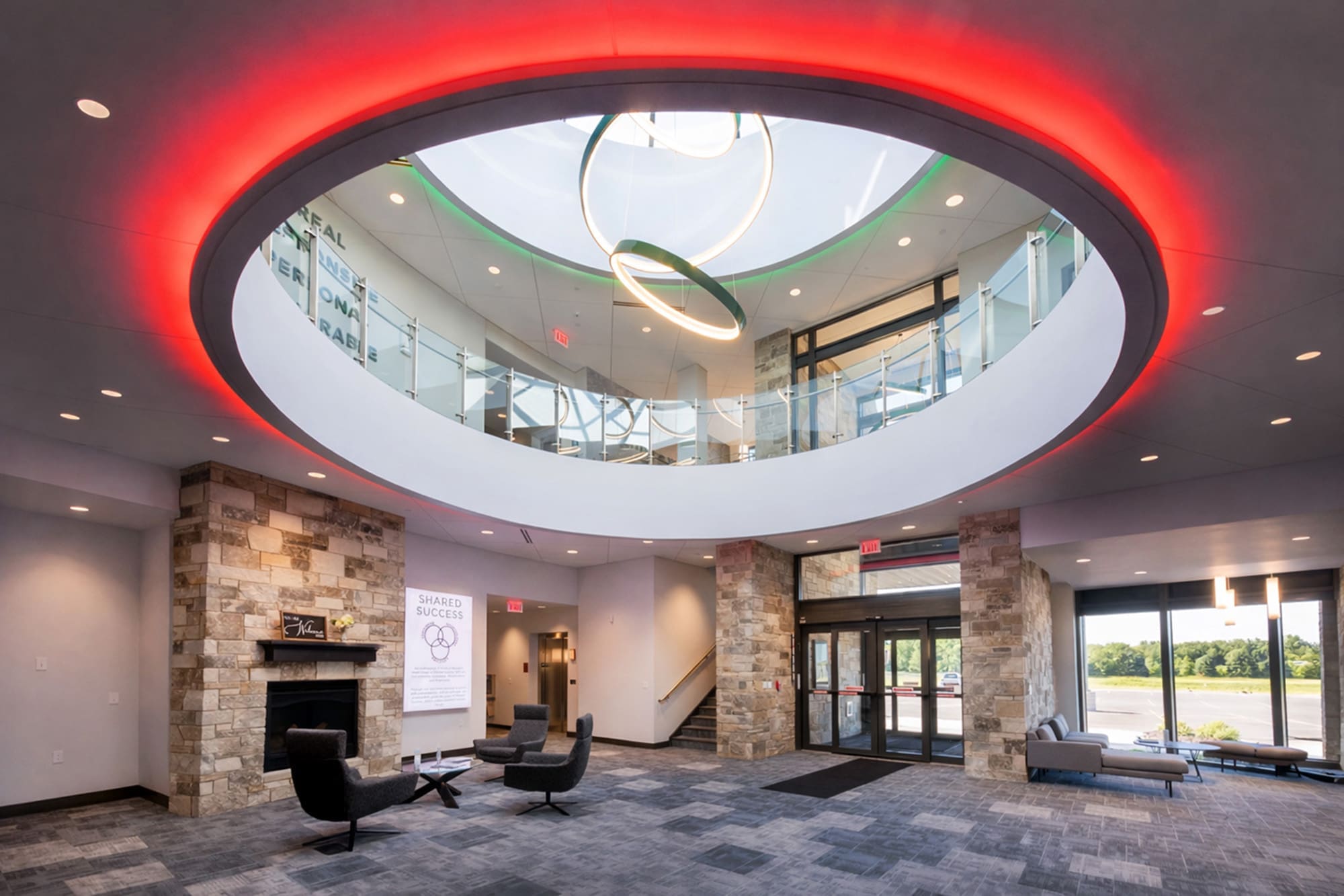 Spacious commercial lobby featuring circular skylight, red ceiling lights, stone fireplace, seating area, large windows, and staircase.