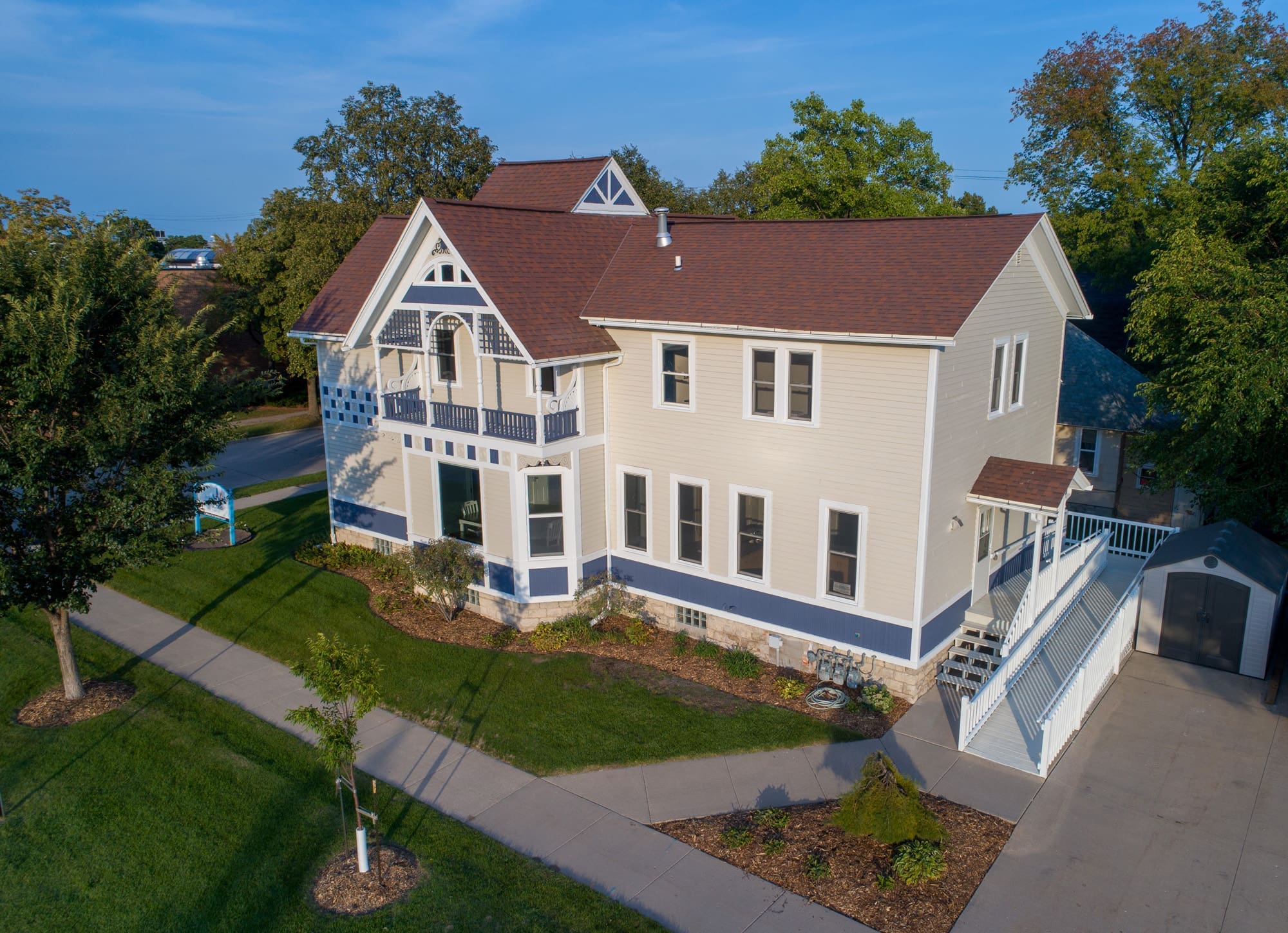 Residential real estate listing photo of a two-story beige home, brown roof, landscaped yard, and concrete walkways in bright sunlight.