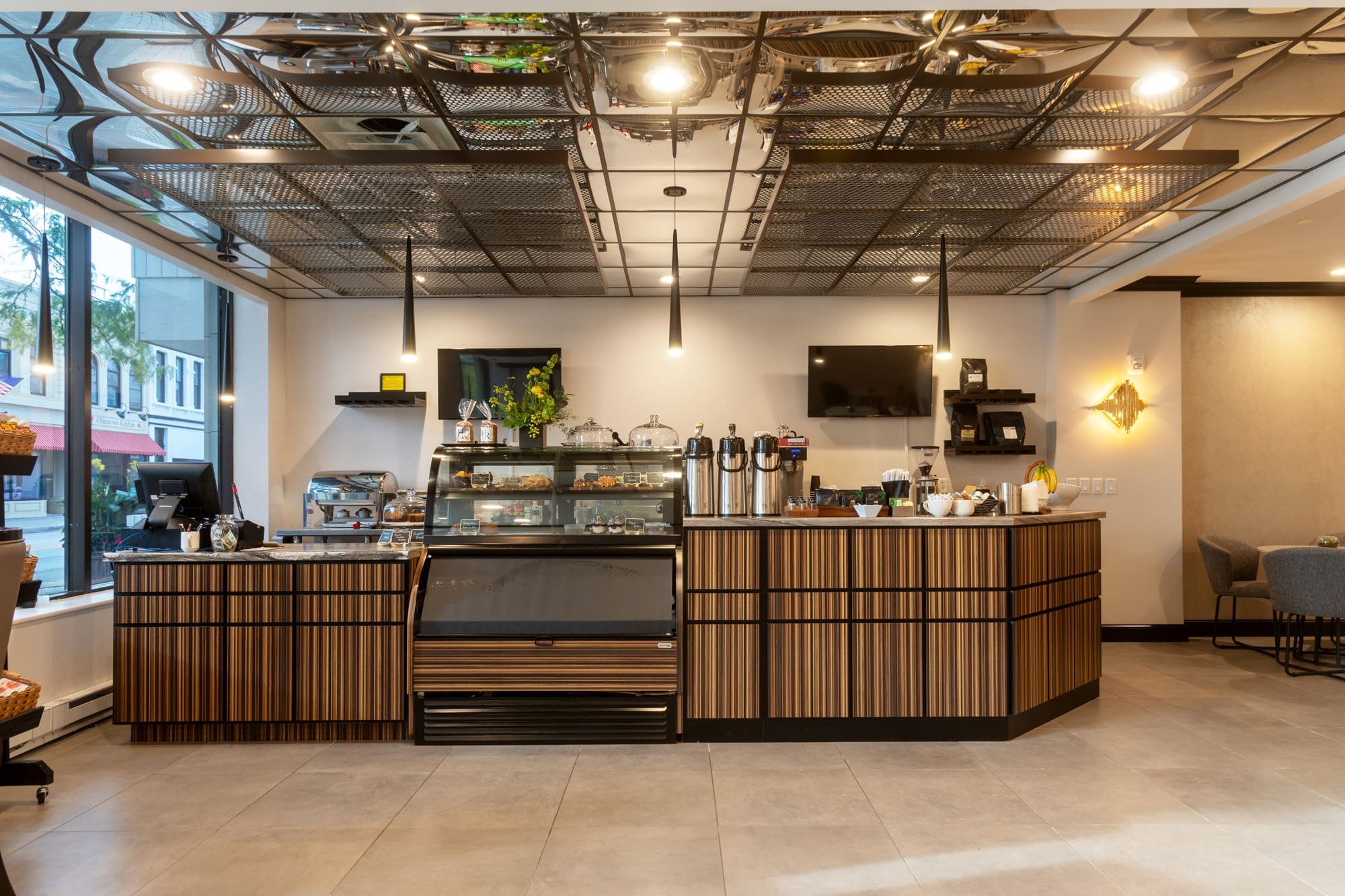 Bright commercial café interior featuring modern counter, pastry case, coffee dispensers, cash register, large windows, and tiled floors.