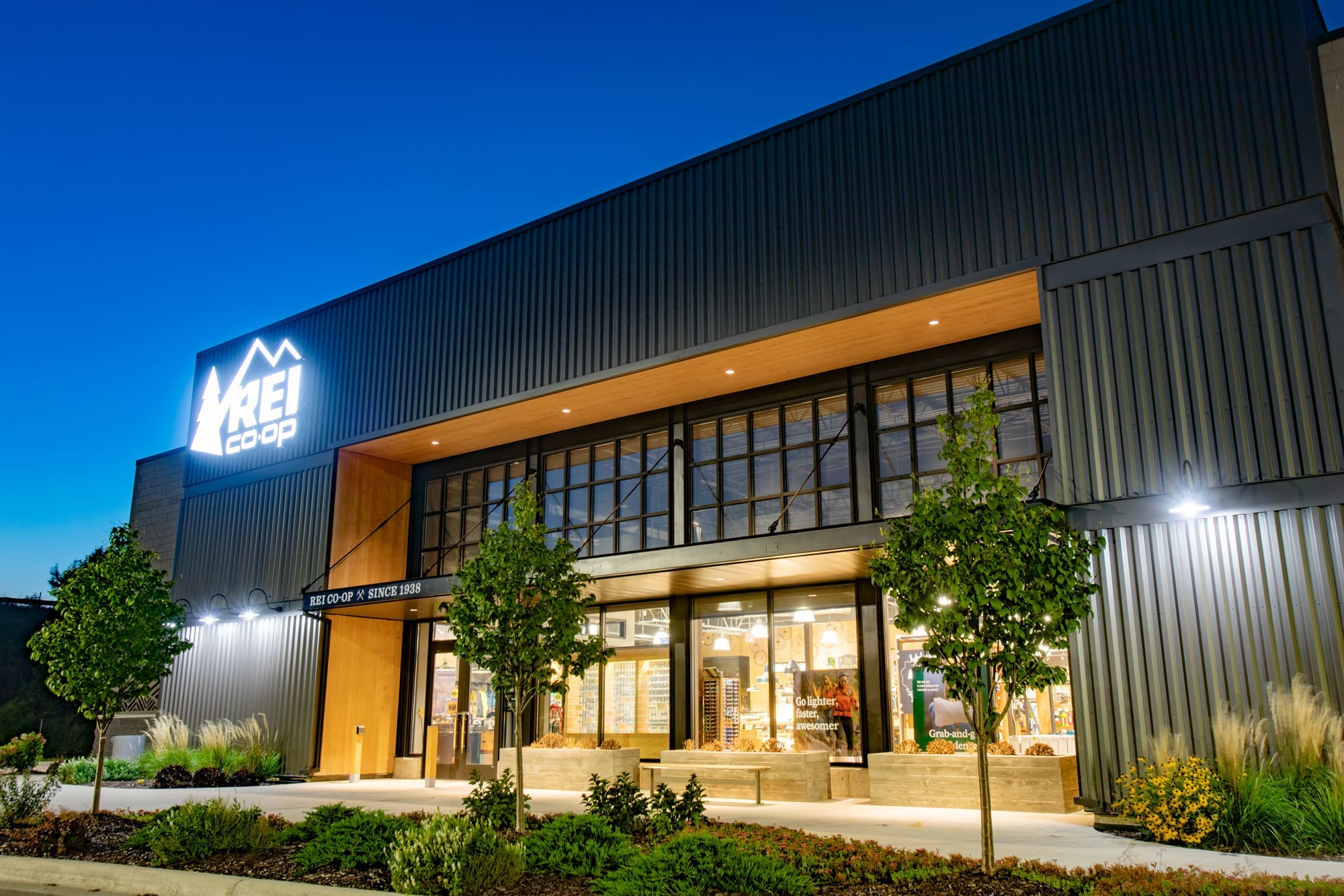 Commercial retail property featuring a modern REI Co-op exterior at dusk, with large windows, lit signage, and landscaped entry.