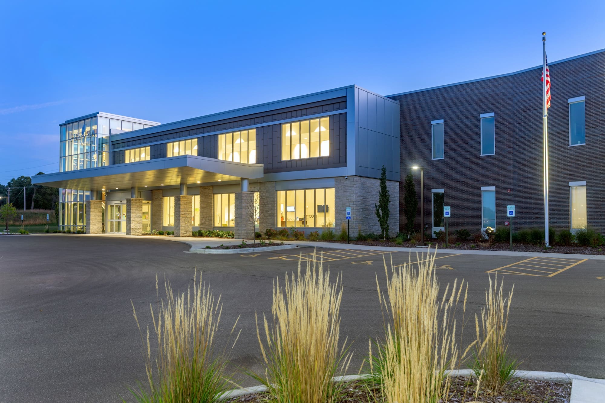 Exterior twilight photo of a modern commercial office building with large windows, entrance canopy, parking, and American flag.