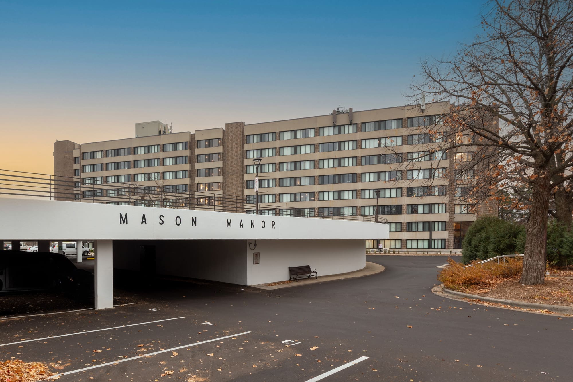 Exterior of Mason Manor, a multi-story residential apartment building with parking garage, driveway, and landscaped autumn trees.