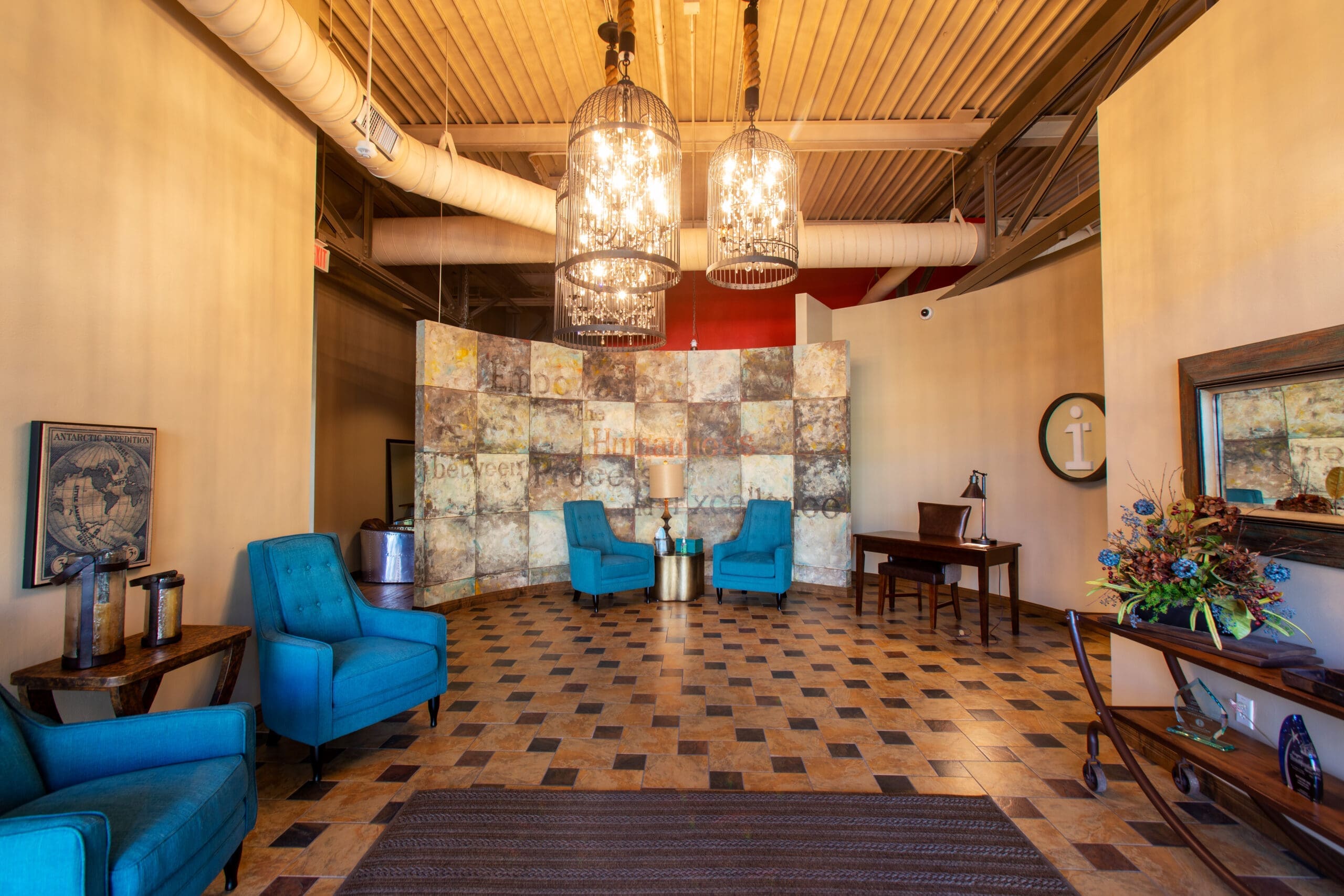 Spacious commercial lobby featuring blue chairs, modern chandeliers, patterned floors, a desk, wall mirror, and decorative plants.