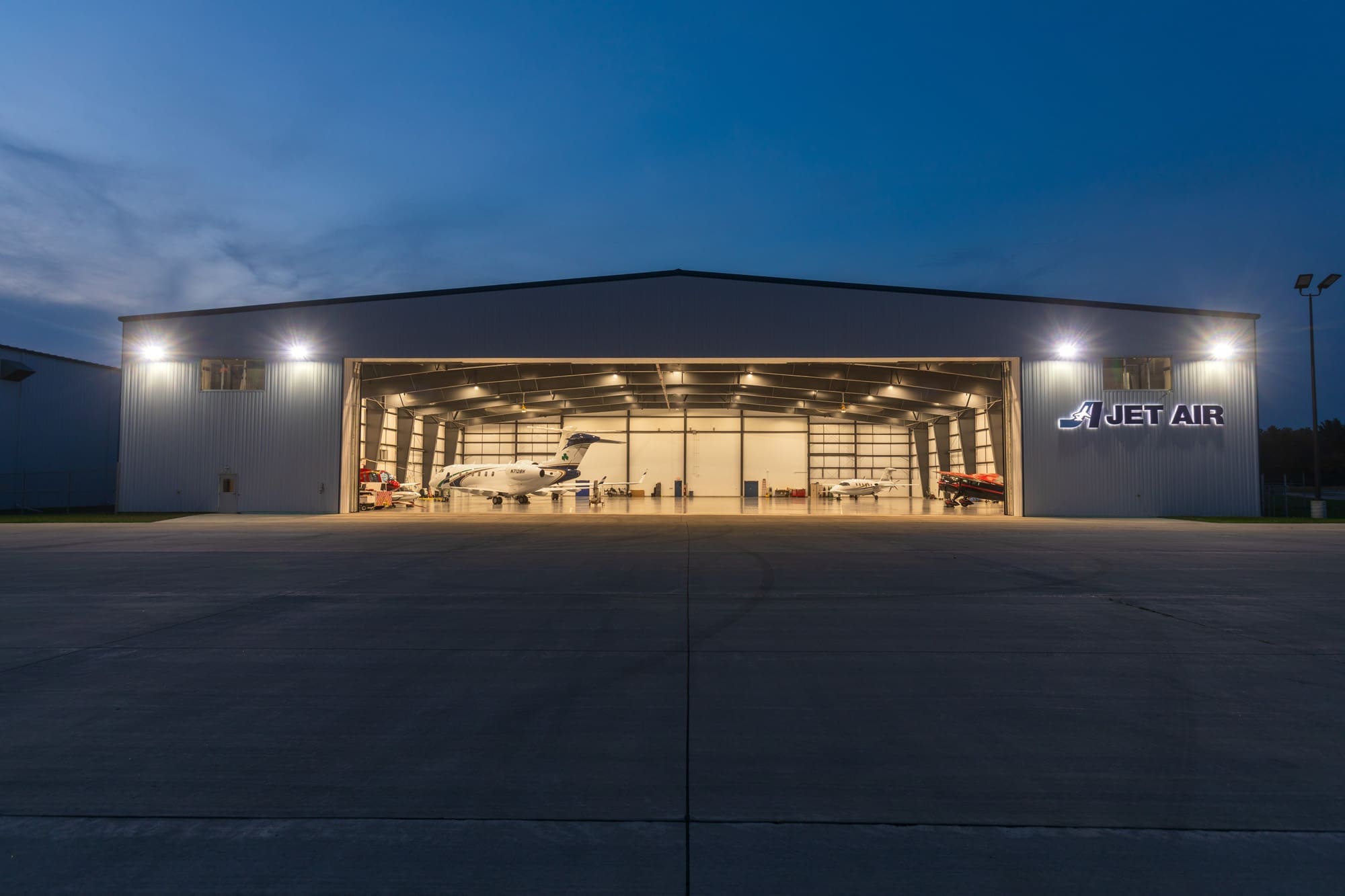 Commercial real estate photo of a well-lit Jet Air airplane hangar at dusk, showcasing spacious interior with aircraft and vehicles.