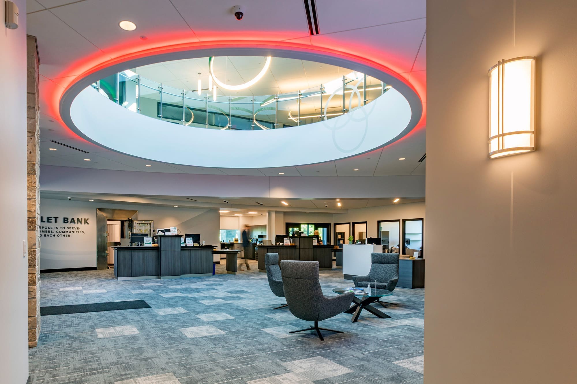 Commercial bank lobby with gray seating, patterned carpet, circular skylight, red accent lighting, and visible office spaces.