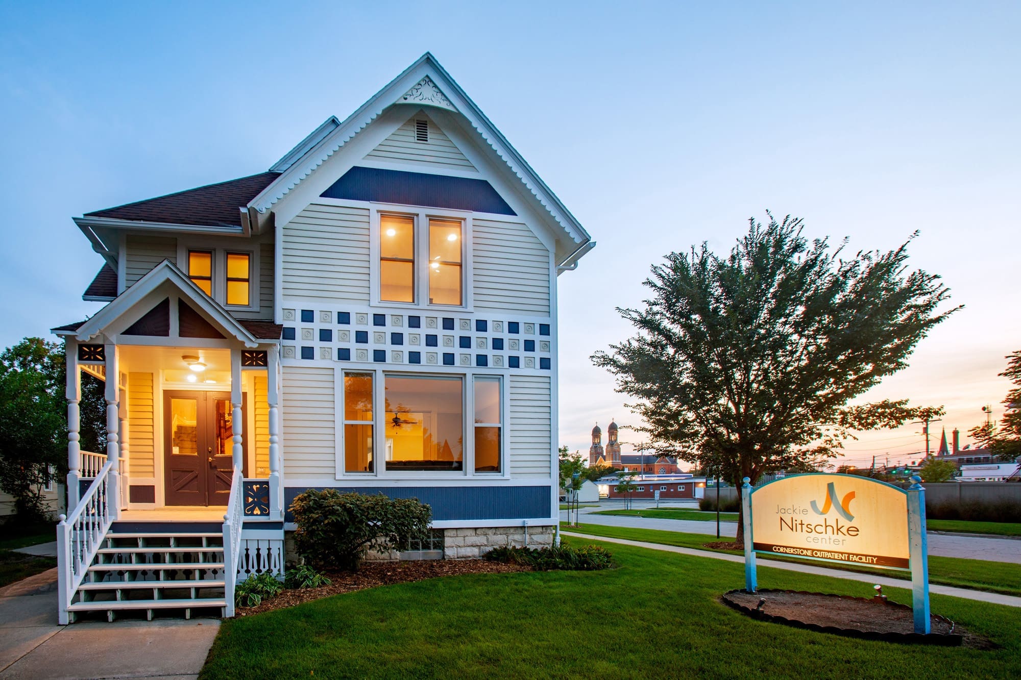 Residential real estate photo of a two-story white and blue home with lit windows at dusk, lawn, street, and “Jack Nitschke Center” sign.