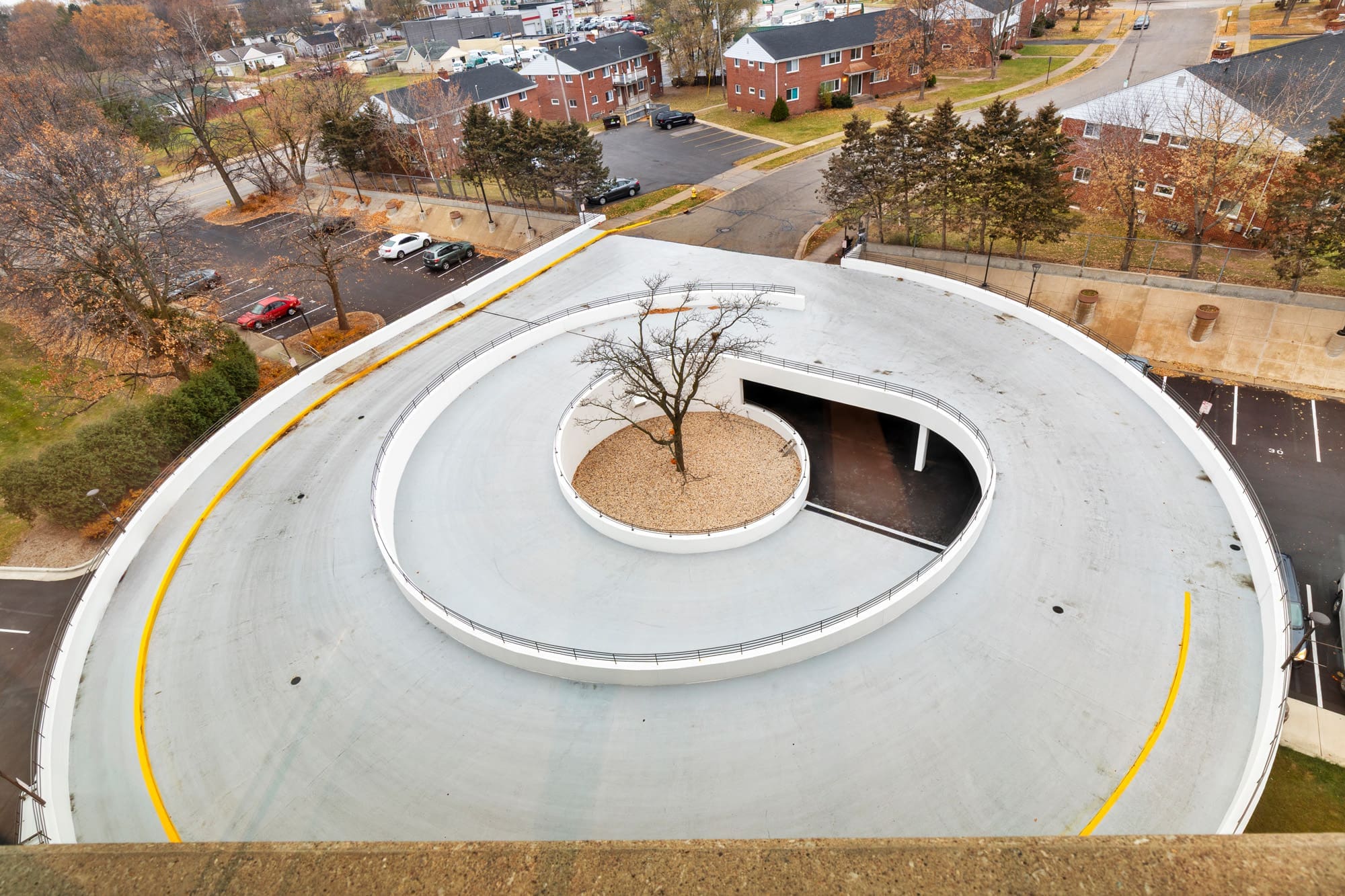 Commercial property exterior featuring a spiral concrete ramp, landscaped tree, adjacent buildings, and parking area in view.
