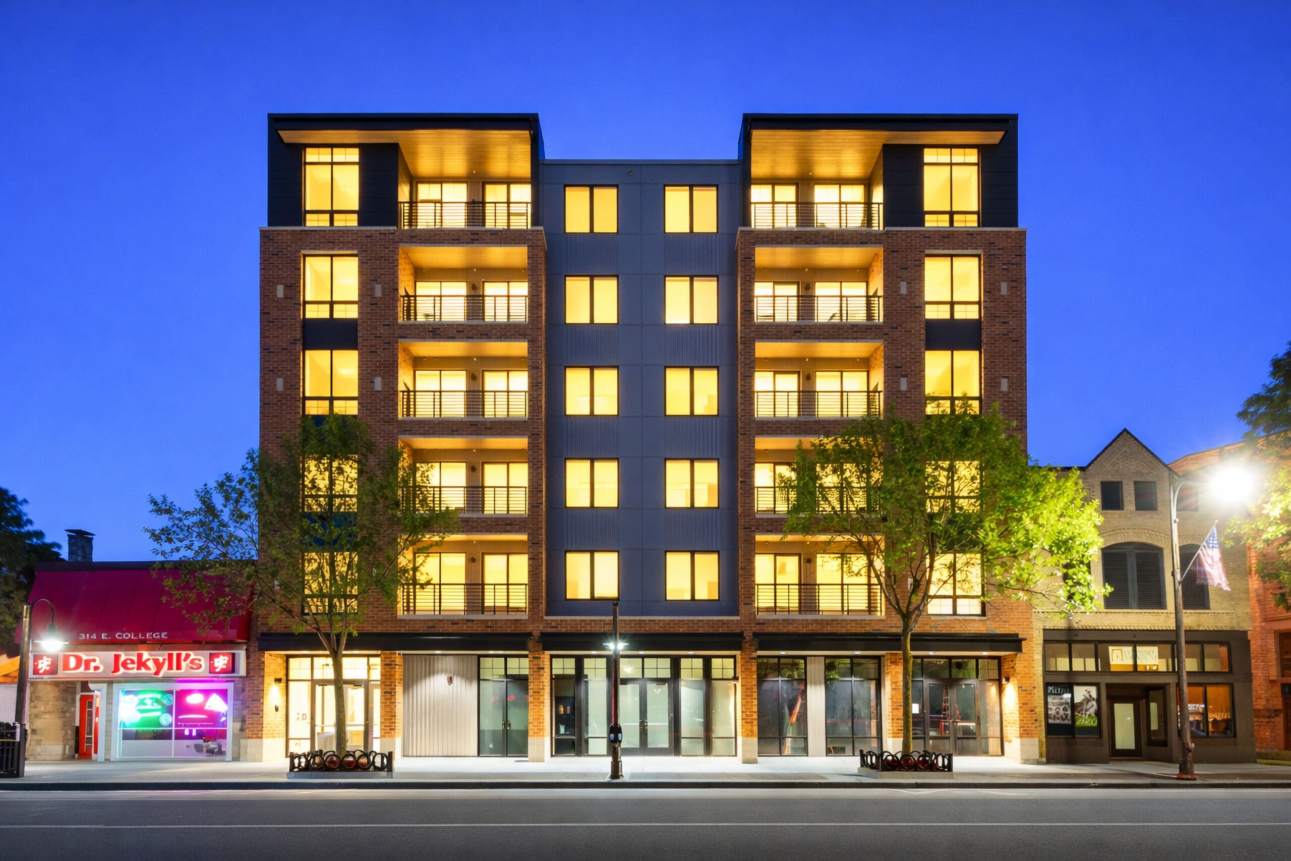 Contemporary mid-rise residential building with glowing windows at dusk, featuring a lit commercial storefront and adjacent historic units.
