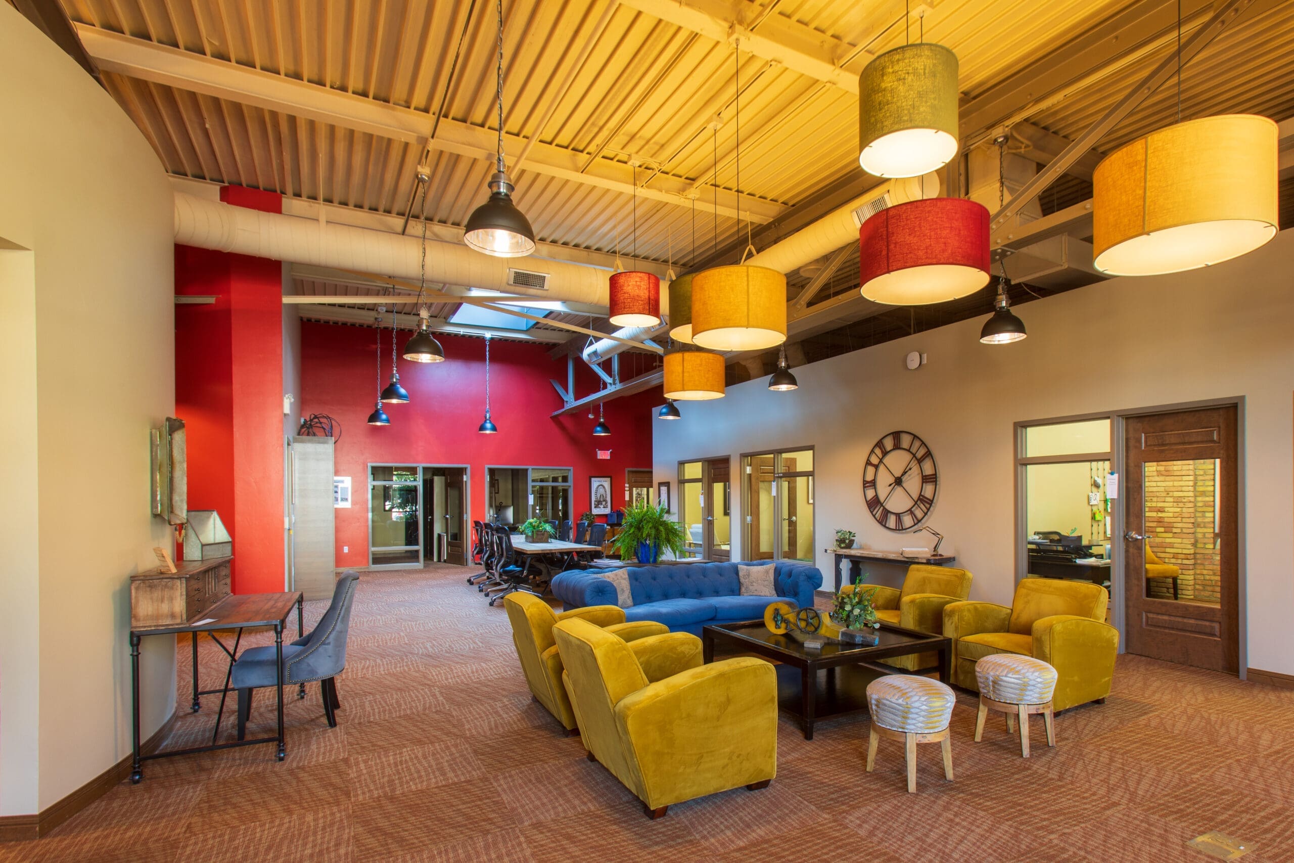 Contemporary commercial office lounge with yellow armchairs, blue sofa, pendant lighting, red accent wall, and glass doors.