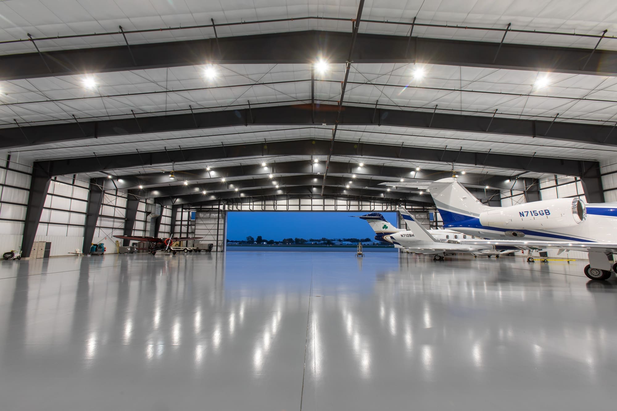 Commercial real estate aircraft hangar with polished floors, ample lighting, multiple private jets, and large doors opening to tarmac.