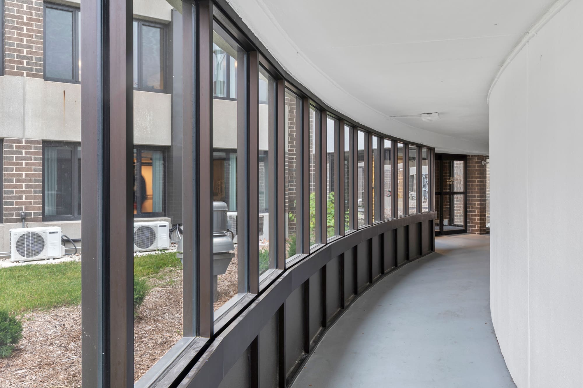 Spacious commercial property hallway featuring large windows with views of landscaped grounds, HVAC units, and adjacent brick building.