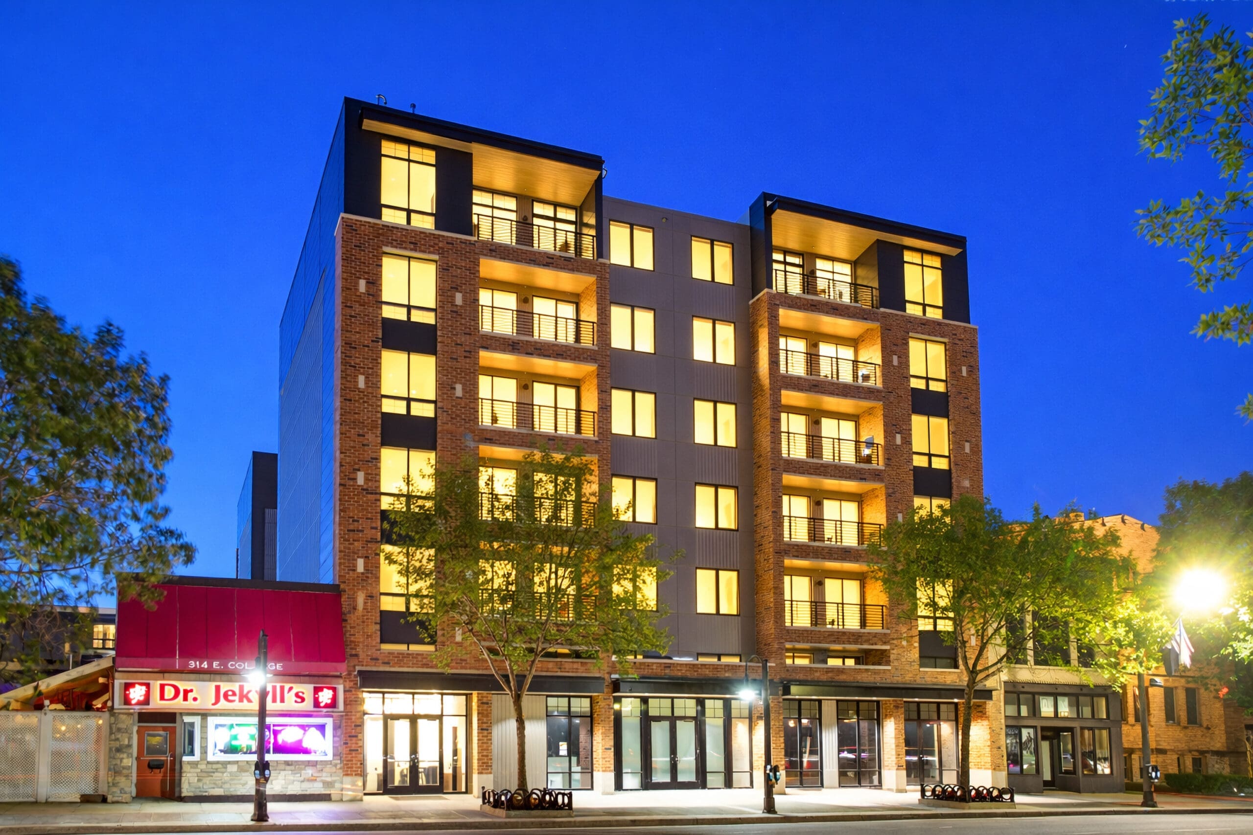 Modern six-story residential apartment building with bright windows beside Dr. Jekylls, ideal real estate listing photo at dusk.