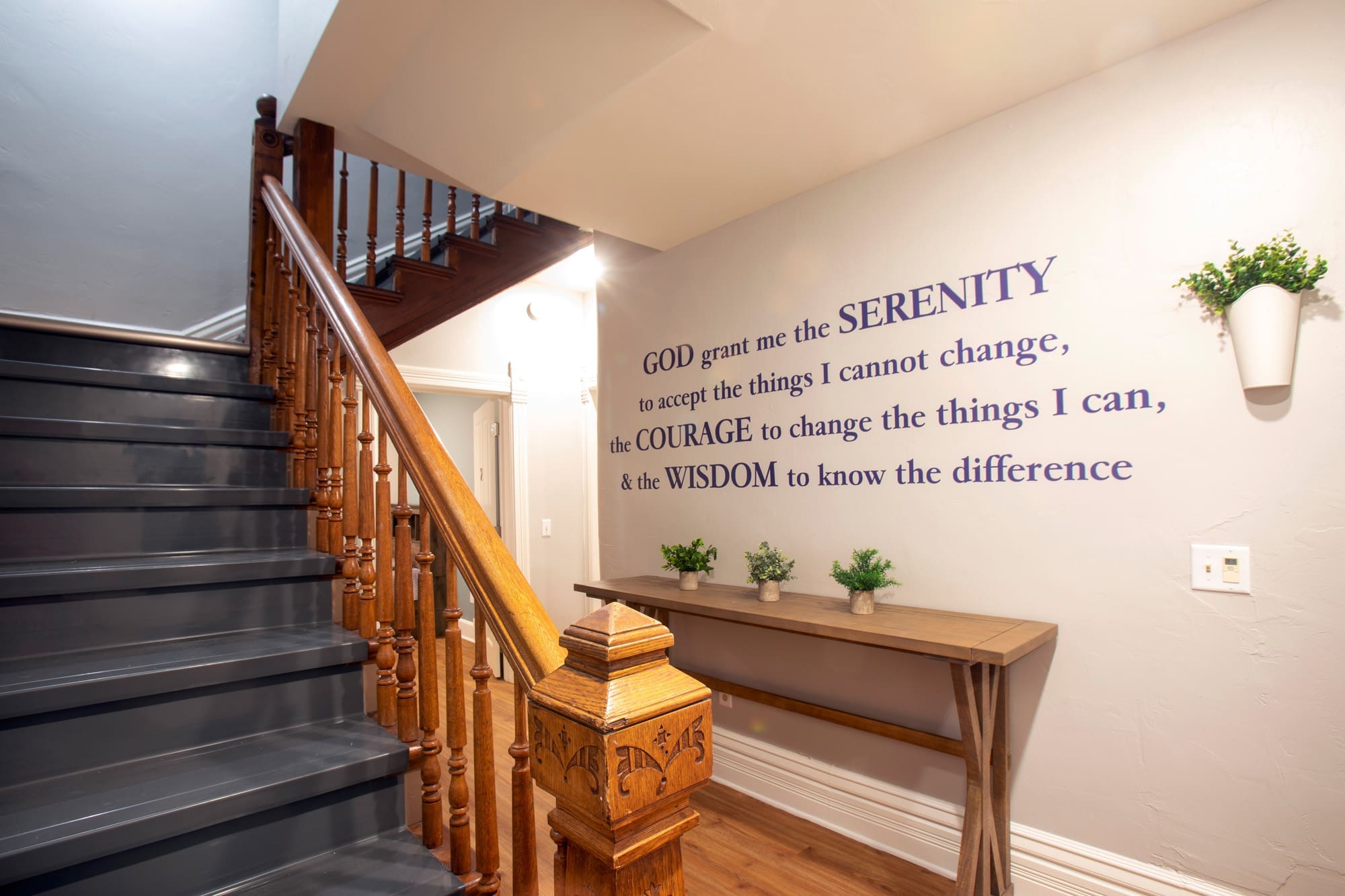 Elegant wooden staircase in residential property, feature wall with Serenity Prayer, console table, and decorative hanging potted plants.