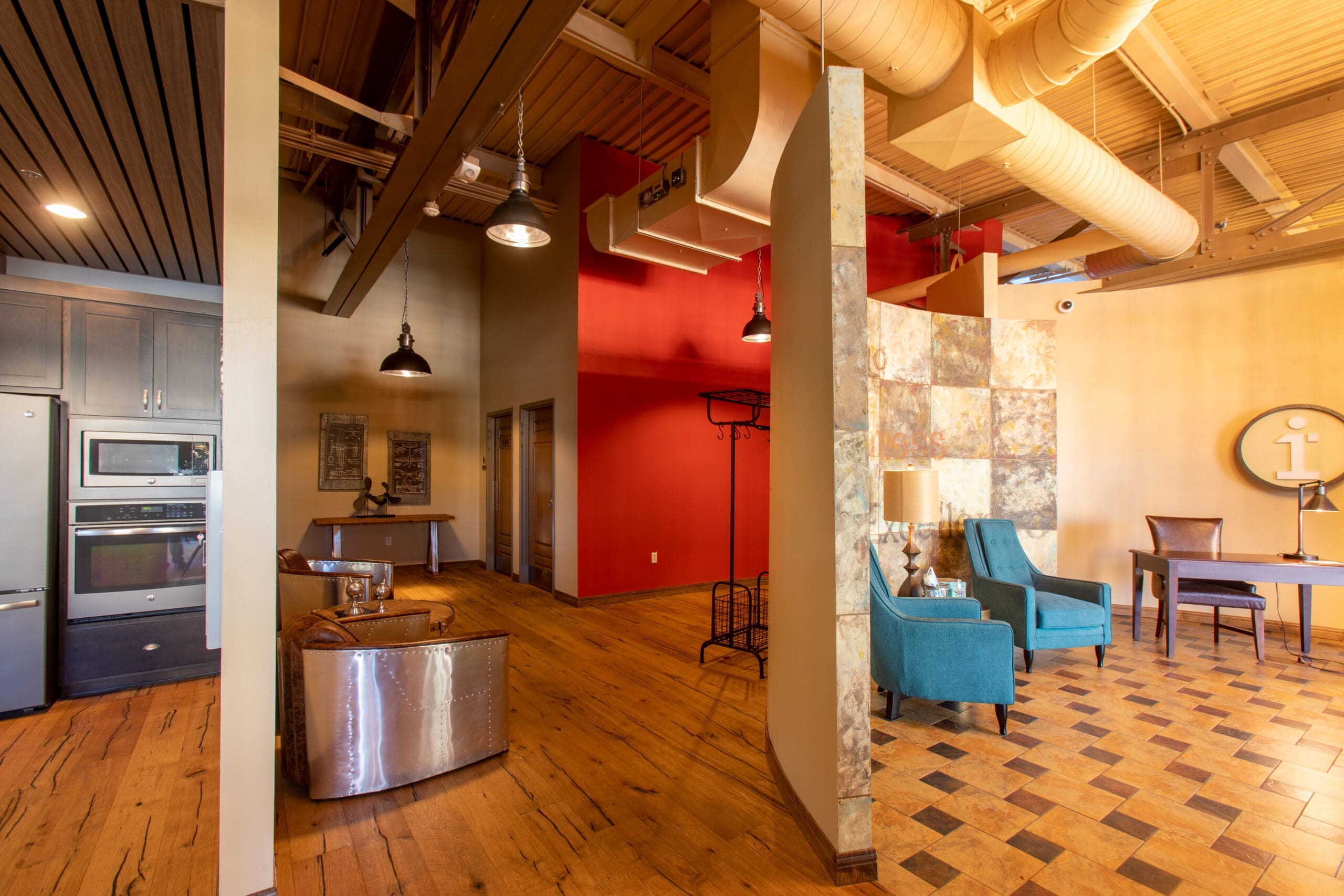 Commercial office space featuring wood floors, blue chairs, metal reception desk, red accent wall, modern lighting, and open ceiling.