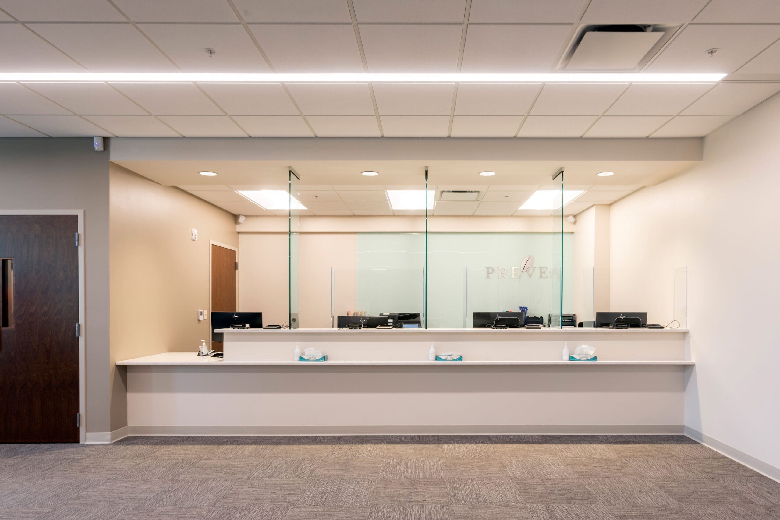 Spacious commercial reception area with modern counter, glass dividers, workstations, neutral walls, and carpeted flooring.