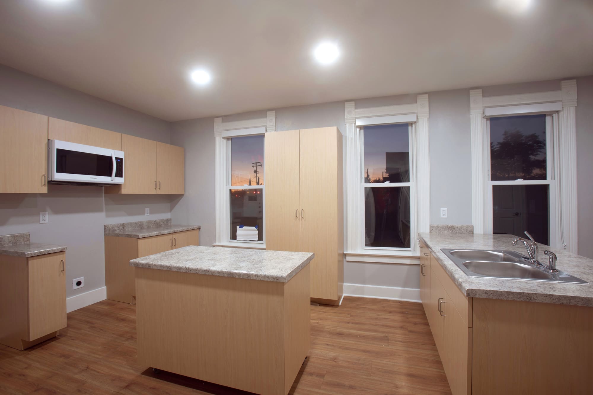Bright residential kitchen with light wood cabinetry, central island, built-in microwave, double sink, and large windows for natural light.