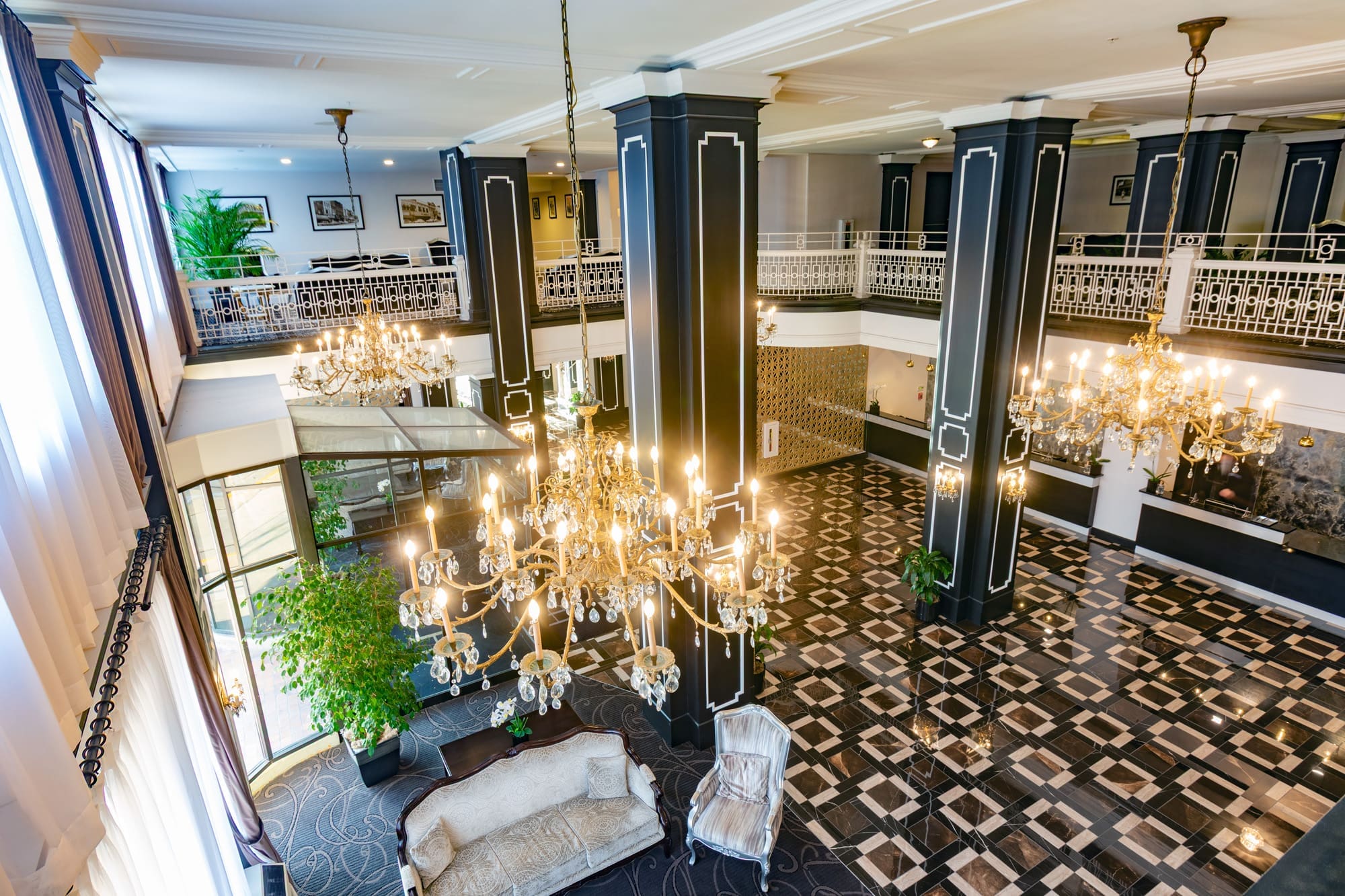 Spacious commercial hotel lobby featuring black and white columns, chandeliers, tile floors, large windows, and mezzanine seating.