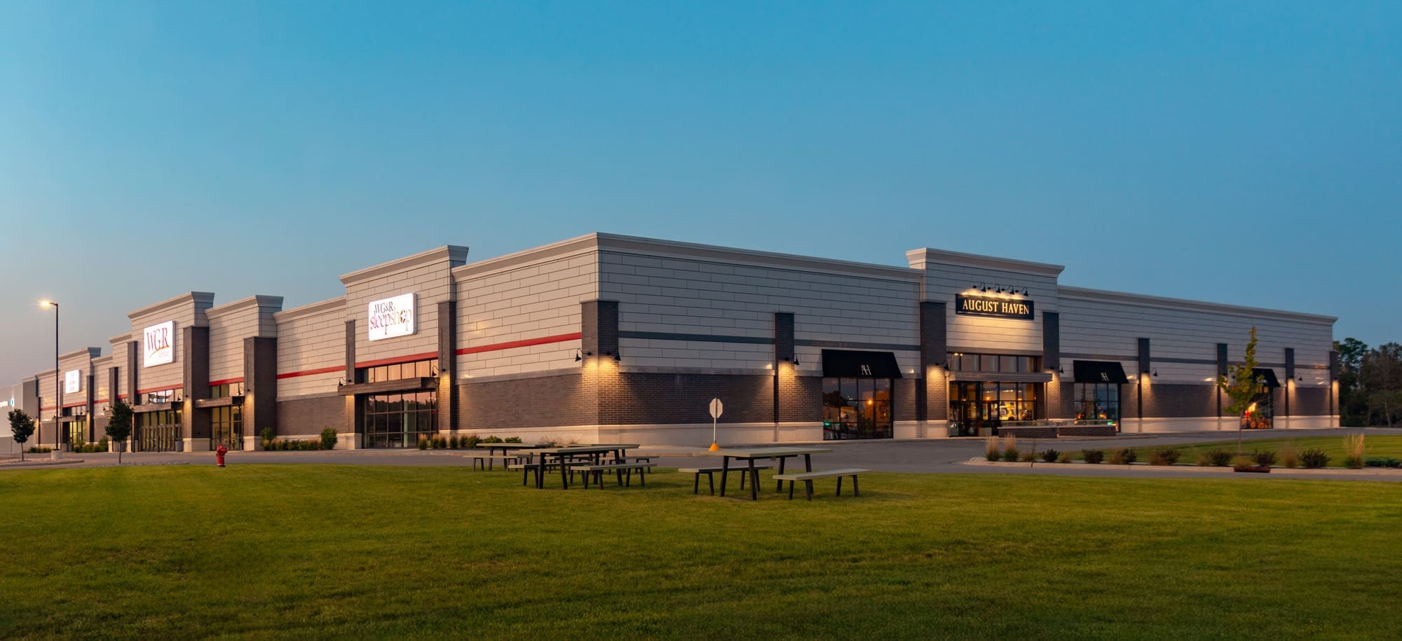 Commercial strip mall property at dusk featuring Academy Sports, multiple storefronts, lawn and picnic tables—ideal real estate listing.