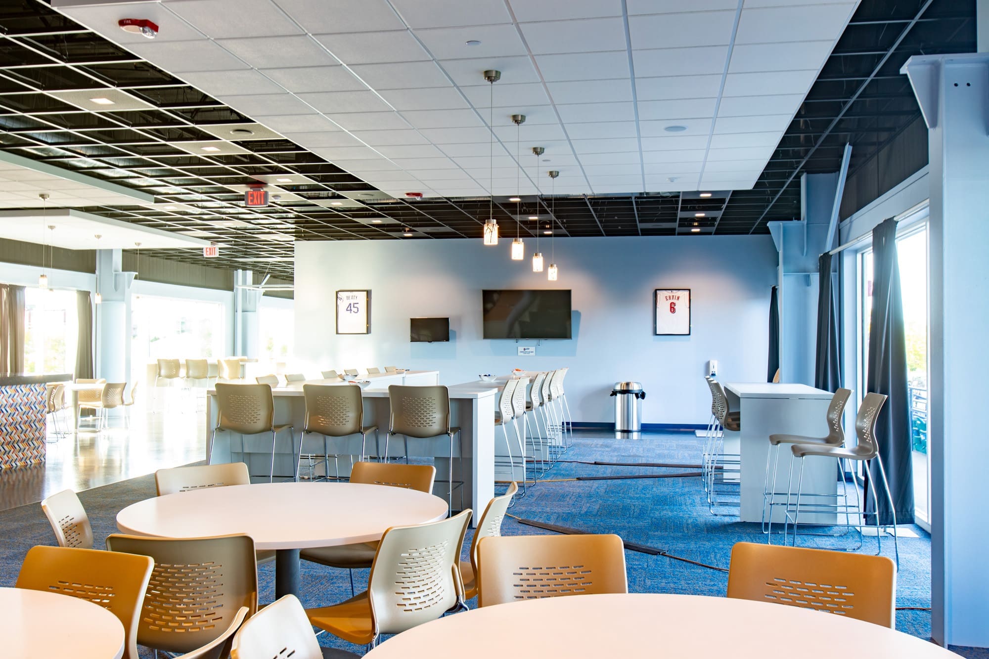 Commercial real estate lounge featuring round tables, bar-height counters, beige chairs, TVs, pendant lighting, and framed jerseys.