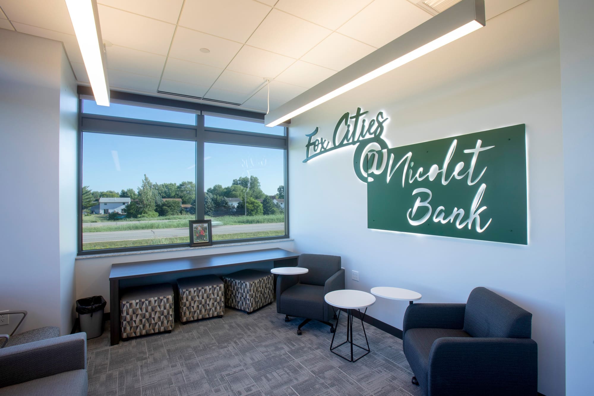 Commercial office lounge featuring gray chairs, small tables, large window with greenery views; signage reads For Cities @Nicolet Bank.