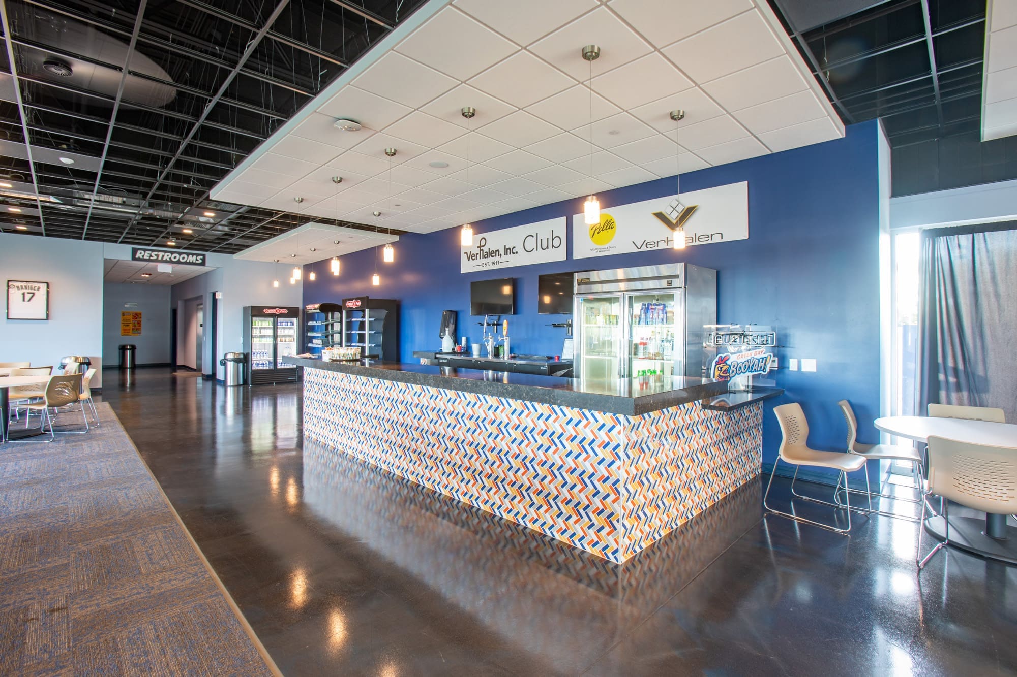 Commercial lounge space with patterned bar counter, blue accent walls, barstools, drink coolers, and club signage.