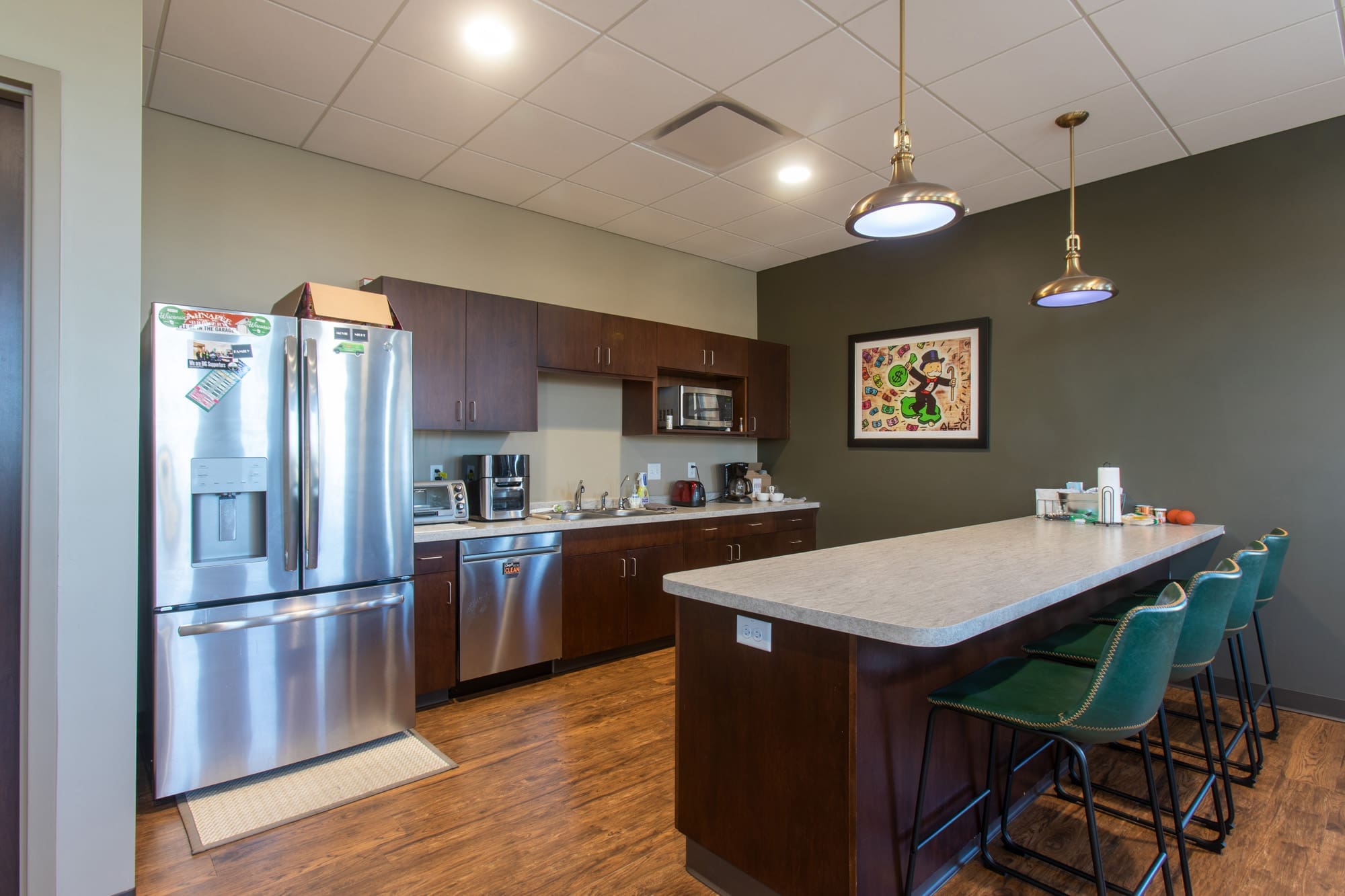 Commercial office kitchen features a stainless steel refrigerator, coffee stations, island counter, green chairs, and modern lighting.