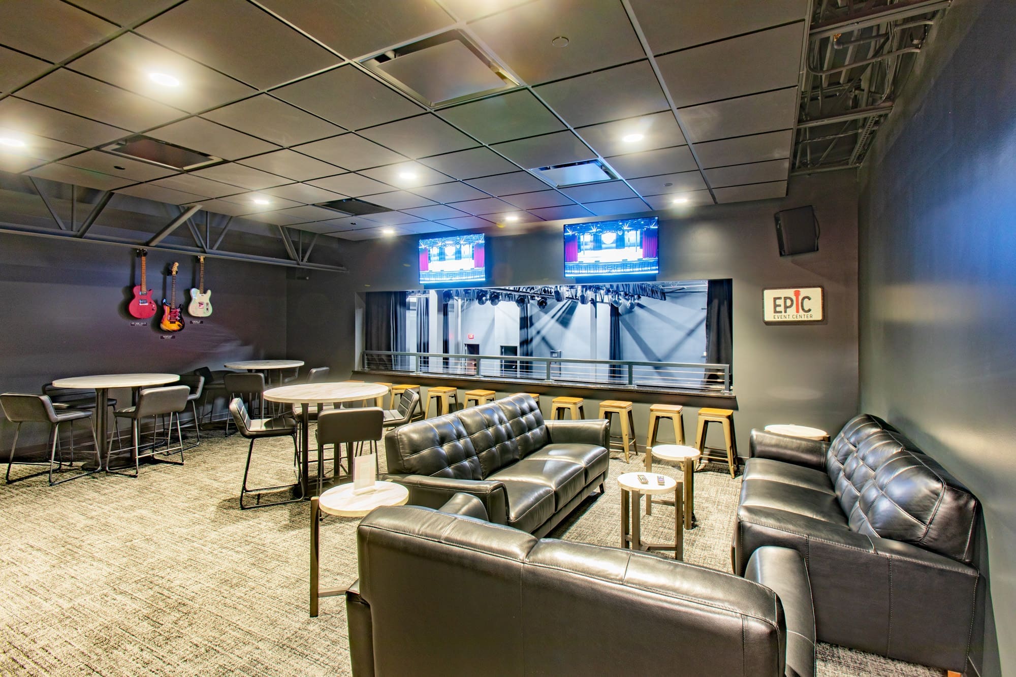 Commercial property lounge featuring black leather sofas, round tables, barstools, mounted guitars, and TVs overlooking stage area.