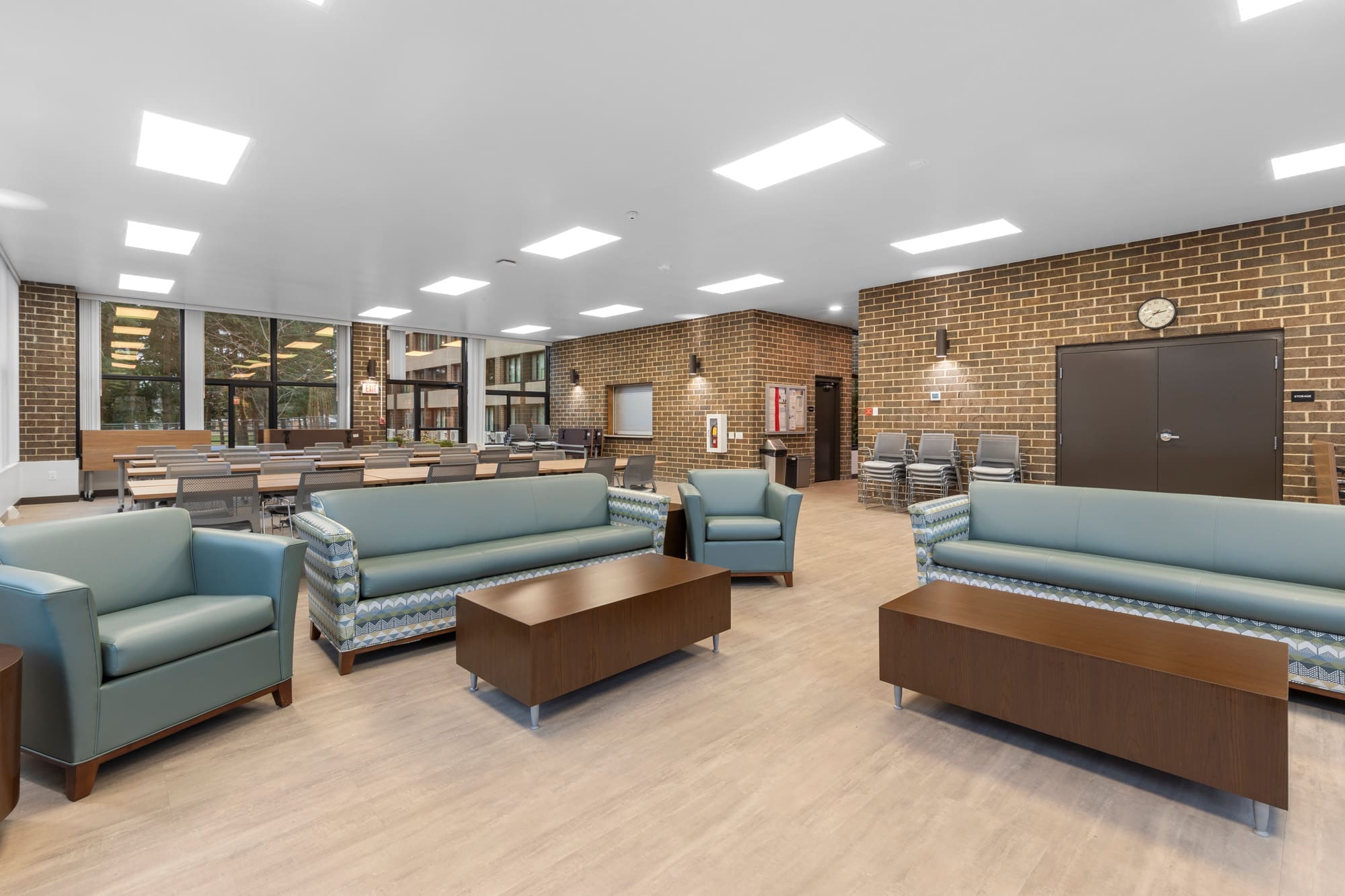 Bright commercial common area featuring modern sofas, armchairs, wood tables, conference setup; brick accent walls, large windows.