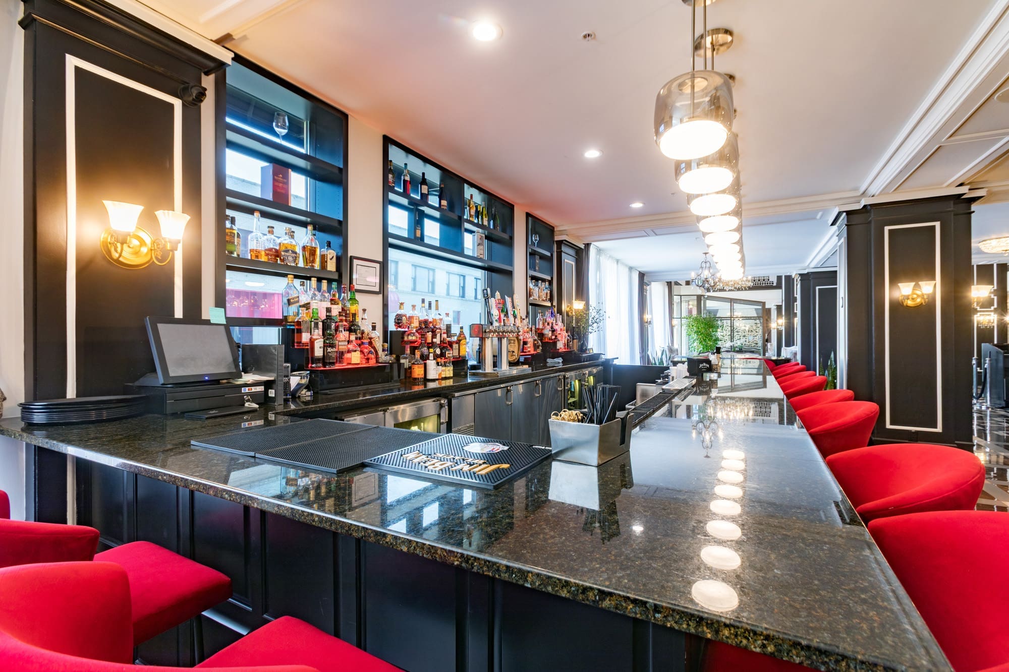 Contemporary commercial bar with a granite countertop, red barstools, liquor display shelves, and pendant lights in a bright space.