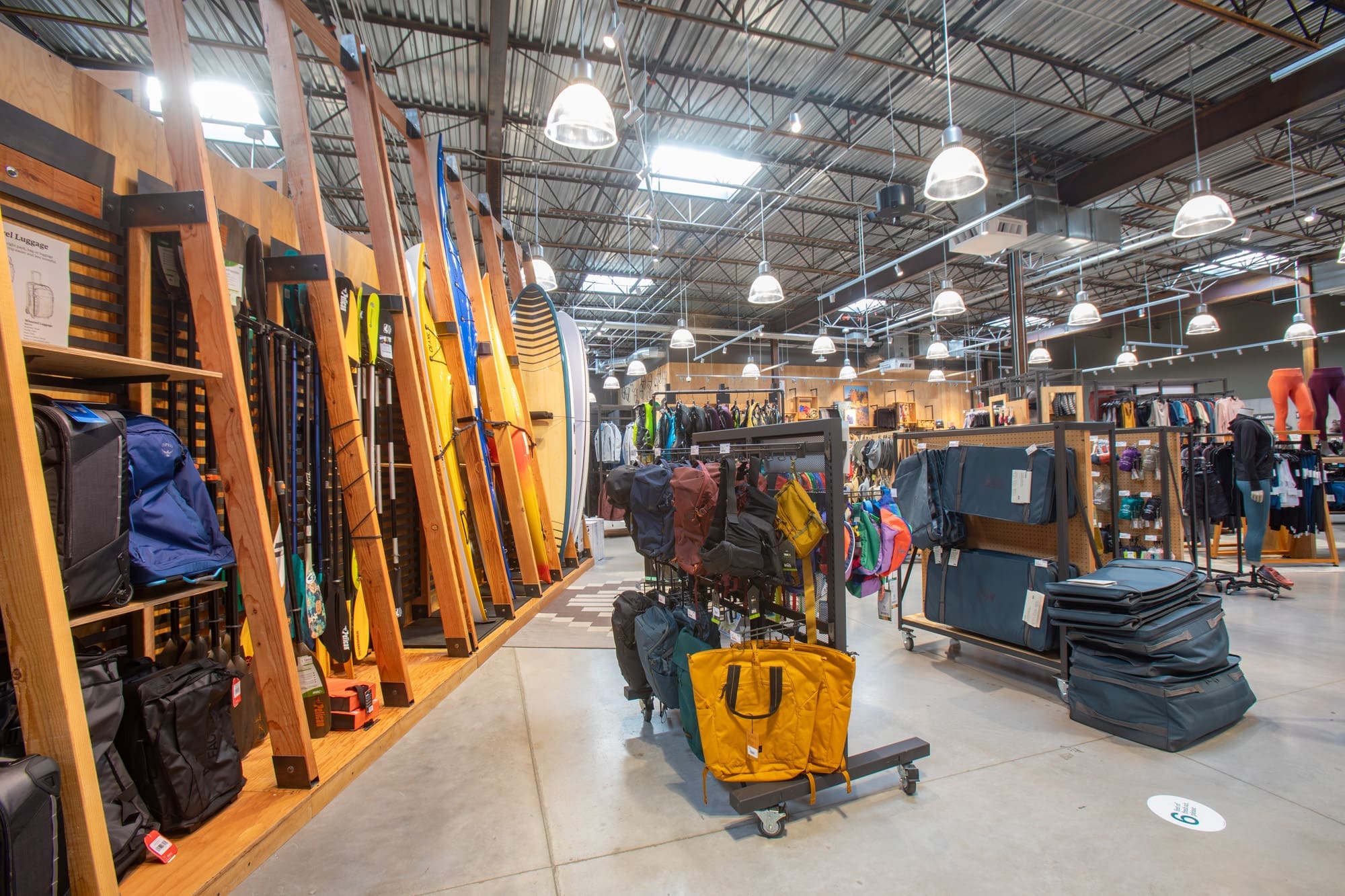 Commercial retail property interior featuring organized displays of outdoor gear, bags, clothing, and paddleboards under modern lighting.