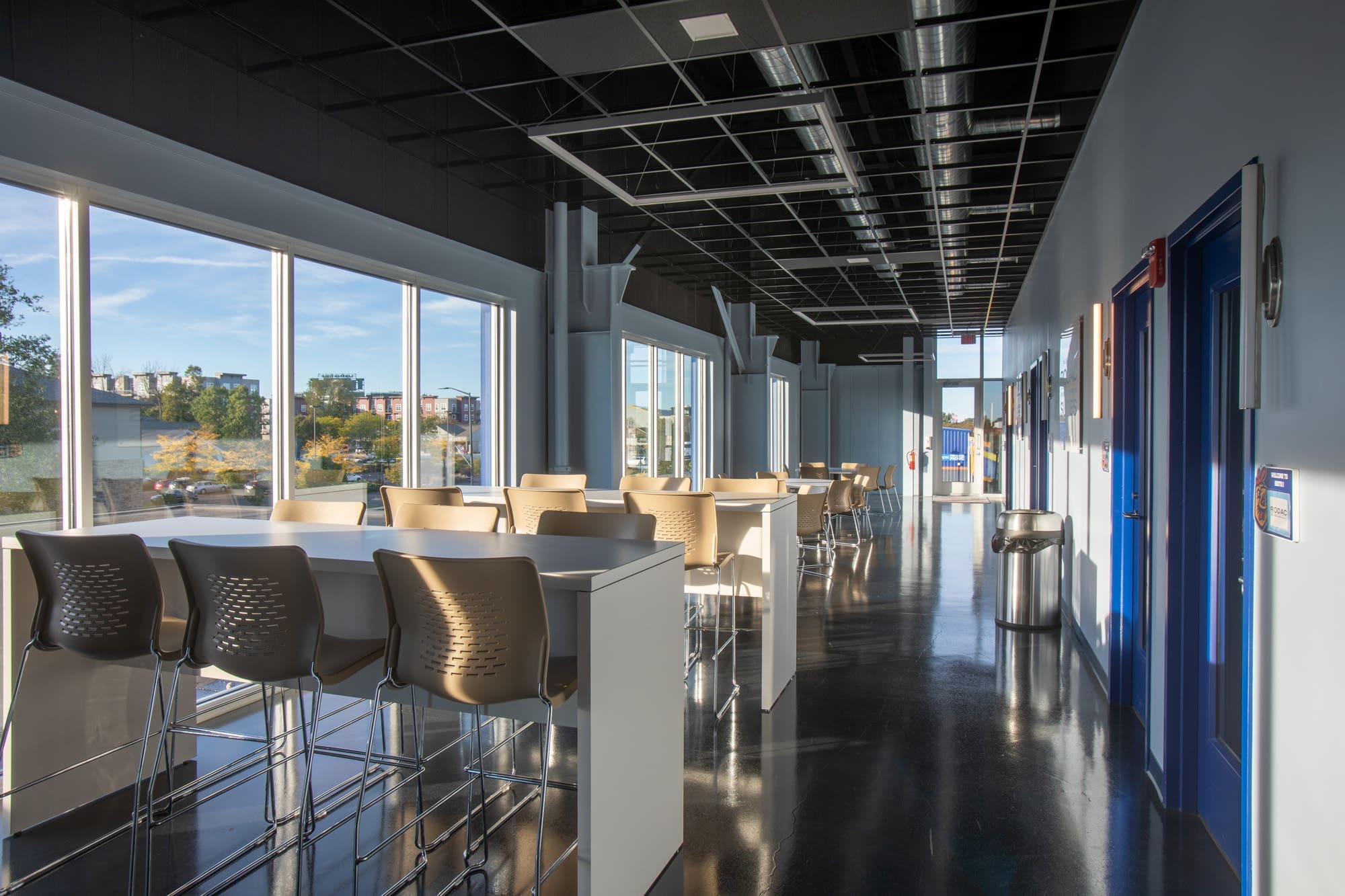 Contemporary commercial corridor with high tables, large windows, glossy floors, blue doors, and abundant natural light.