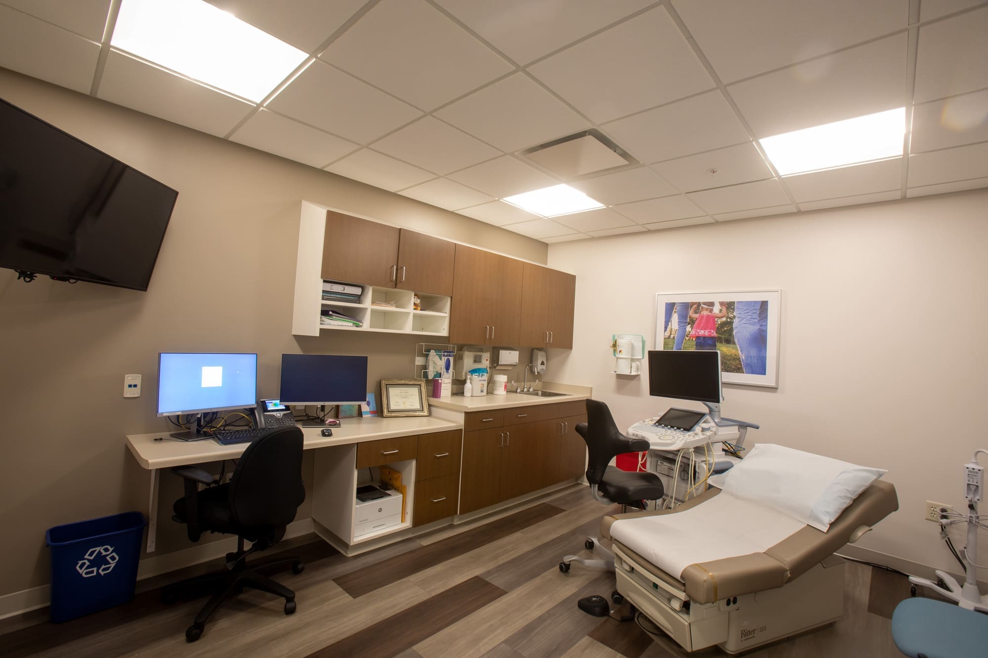 Modern commercial medical exam room featuring an exam table, workstations, cabinetry, equipment, and wall-mounted TV—ideal for clinics.