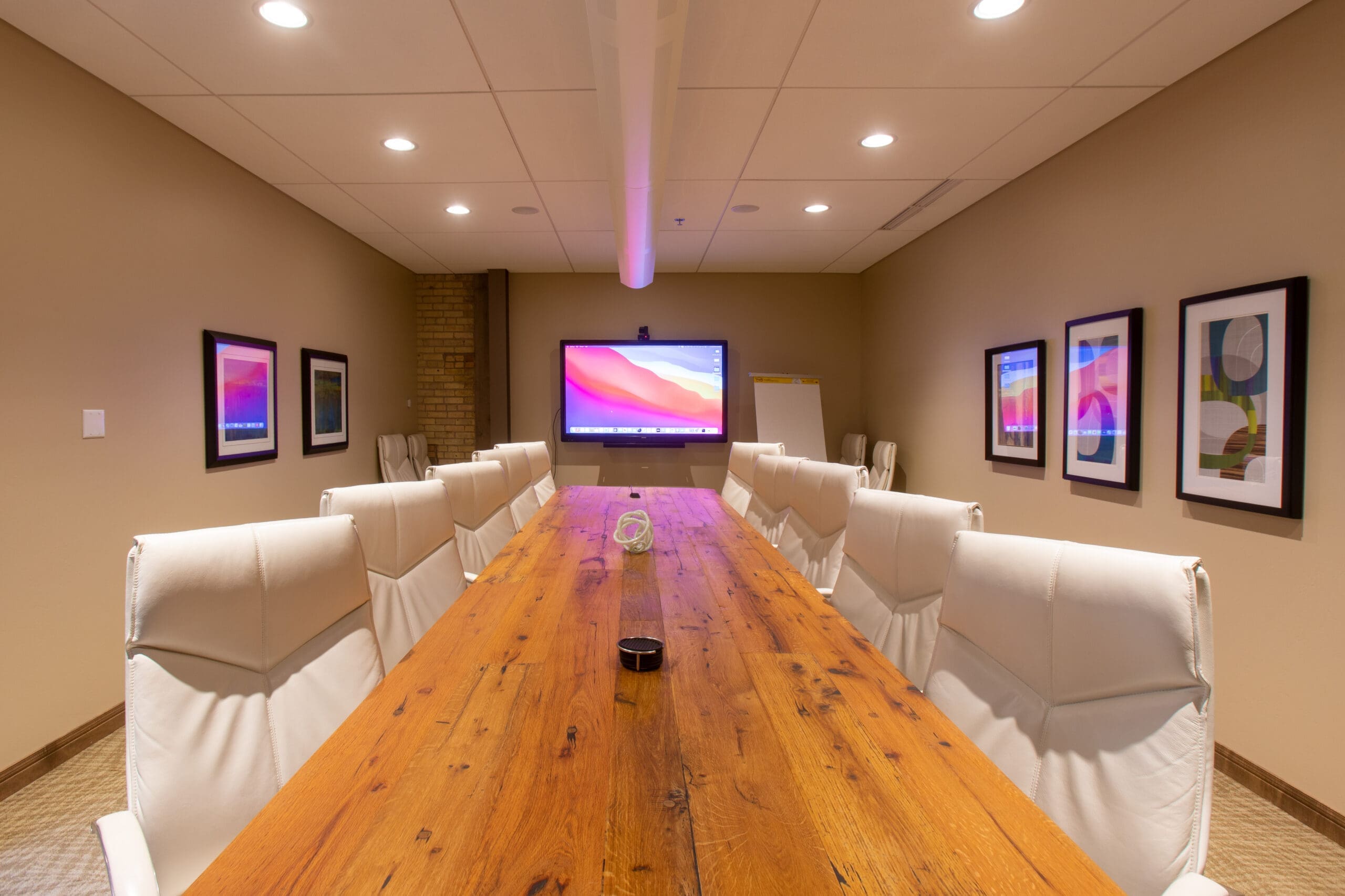 Modern commercial office space featuring a long wooden conference table, white chairs, wall-mounted screen, framed art, and flip chart.