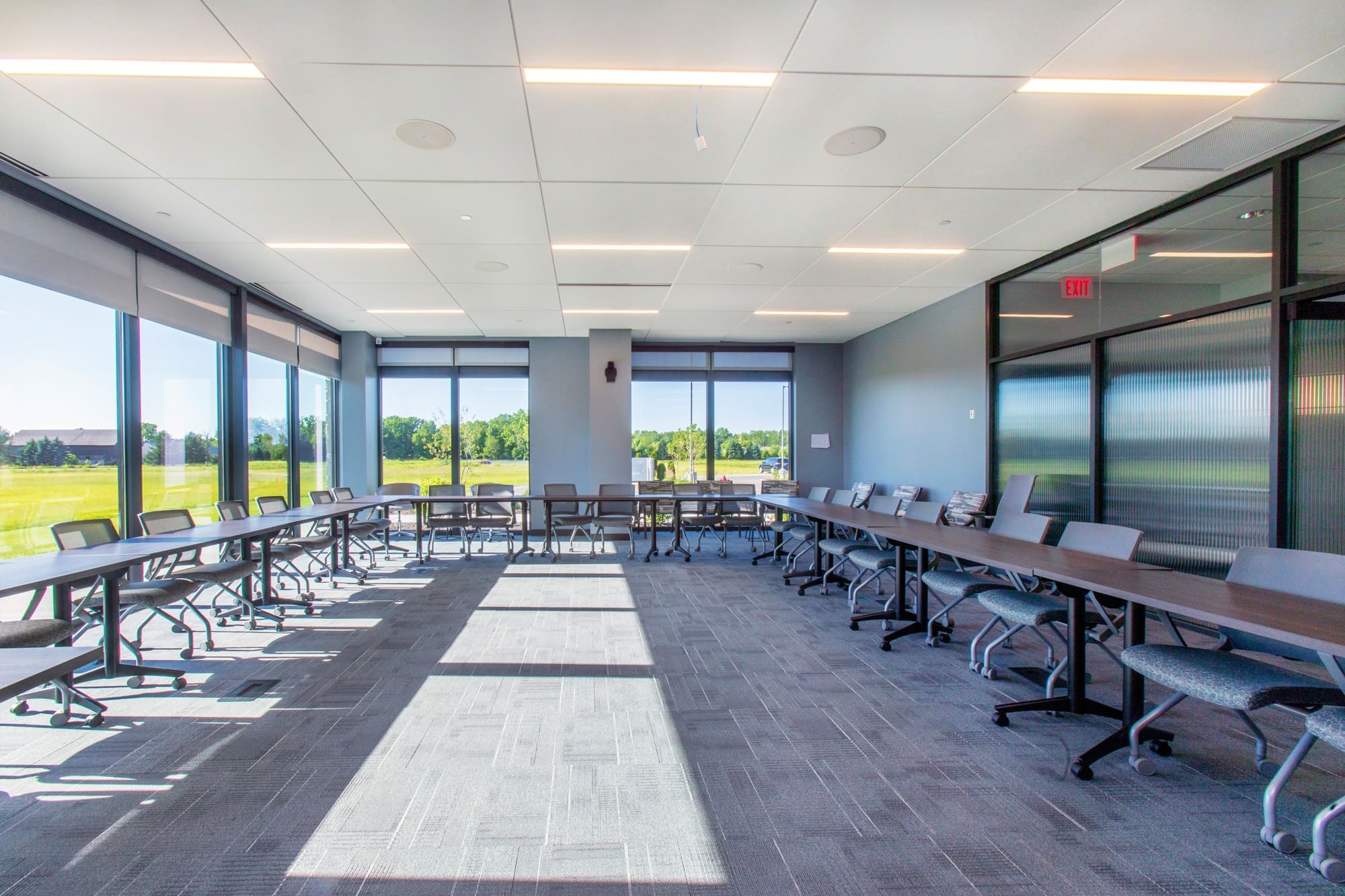 Modern commercial conference room with large windows, abundant natural light, gray carpet, and U-shaped tables with office chairs.