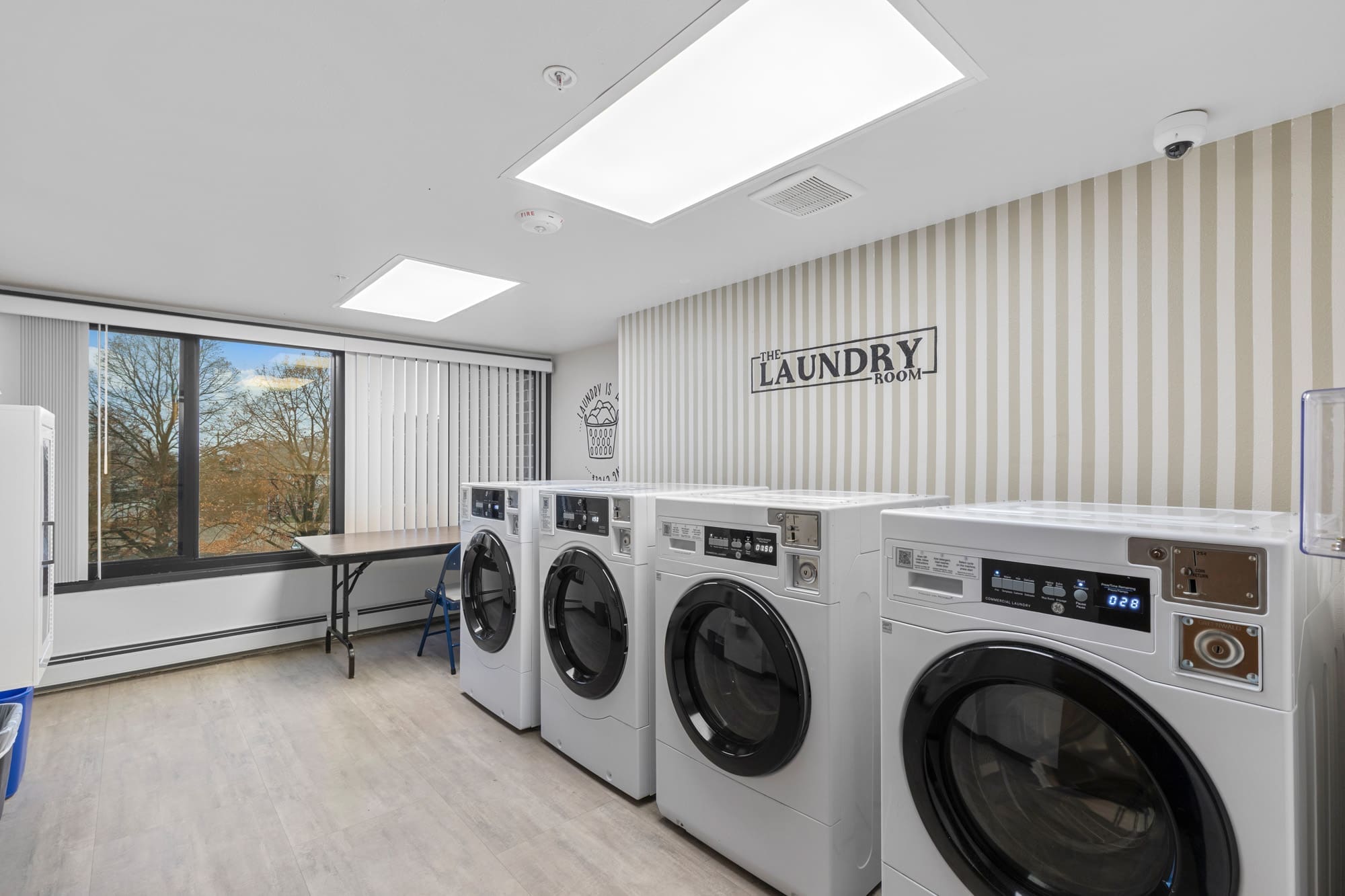 Modern residential laundry room featuring four front-load washers and dryers, folding table, striped wallpaper, and Laundry Room sign.