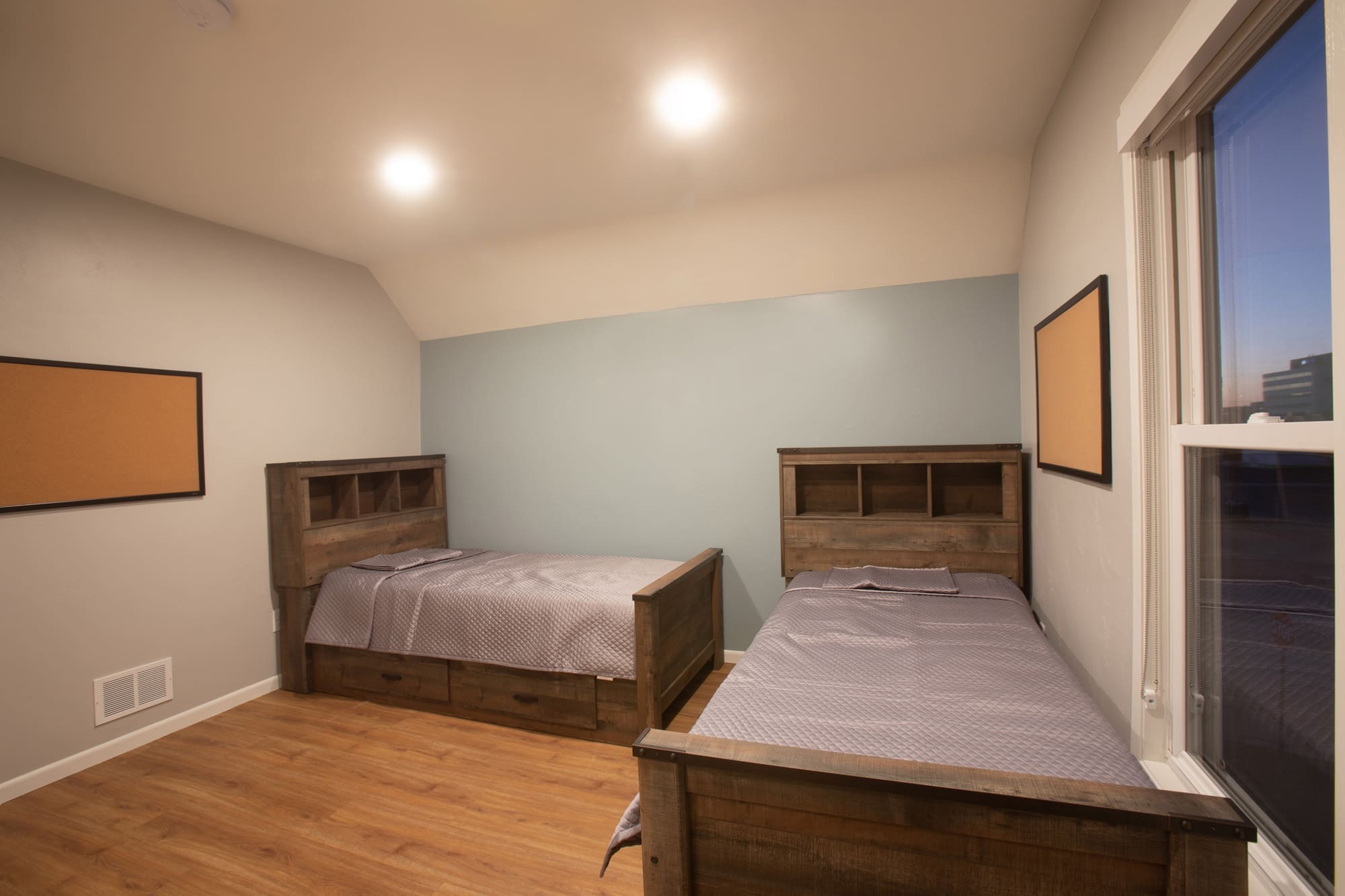 Residential bedroom with two twin beds, headboard shelves, gray bedding, hardwood floors, window, and light-colored walls.