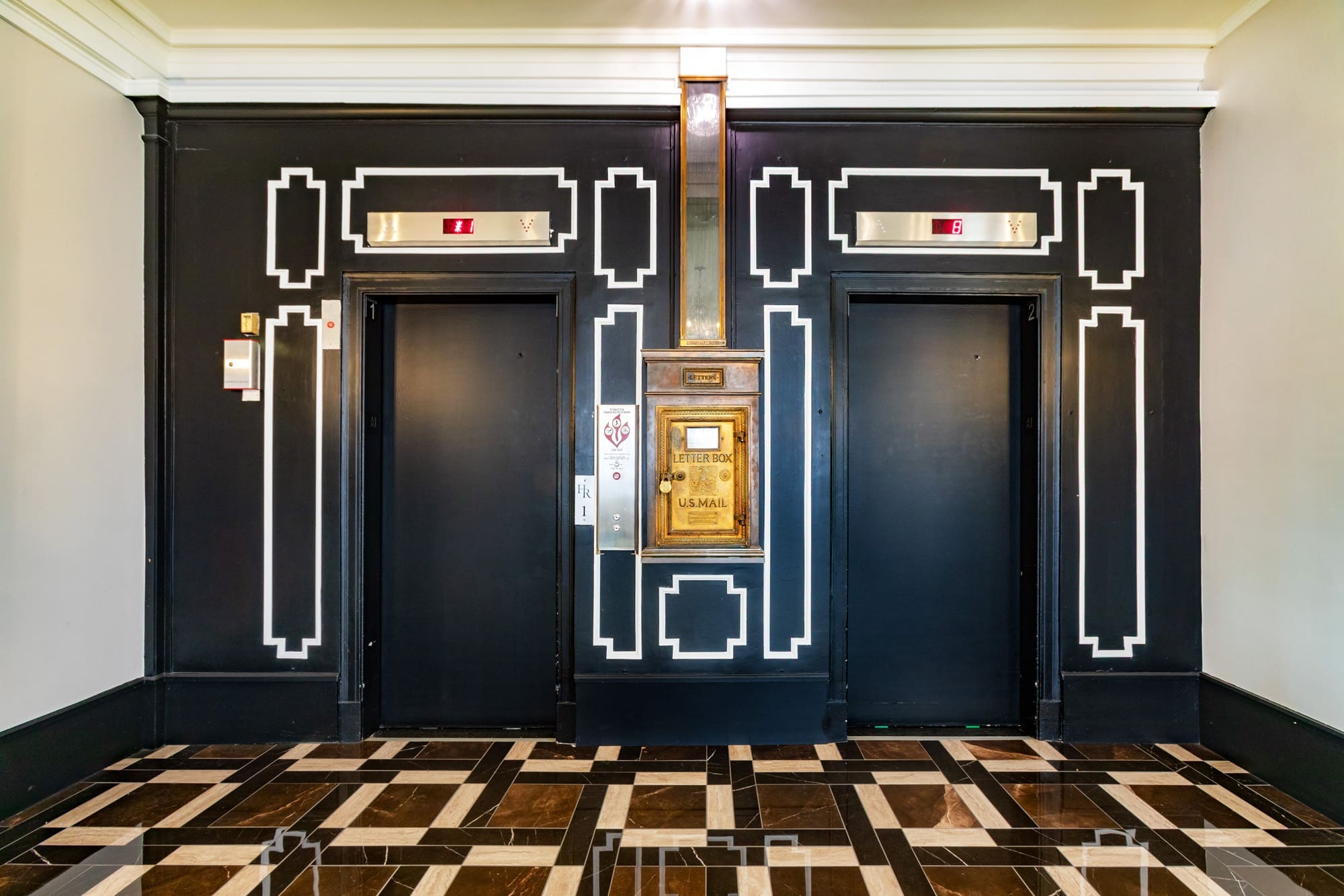 Elegant commercial lobby with black elevator doors, white trim, brass mail chute, and modern black-and-white geometric tile flooring.