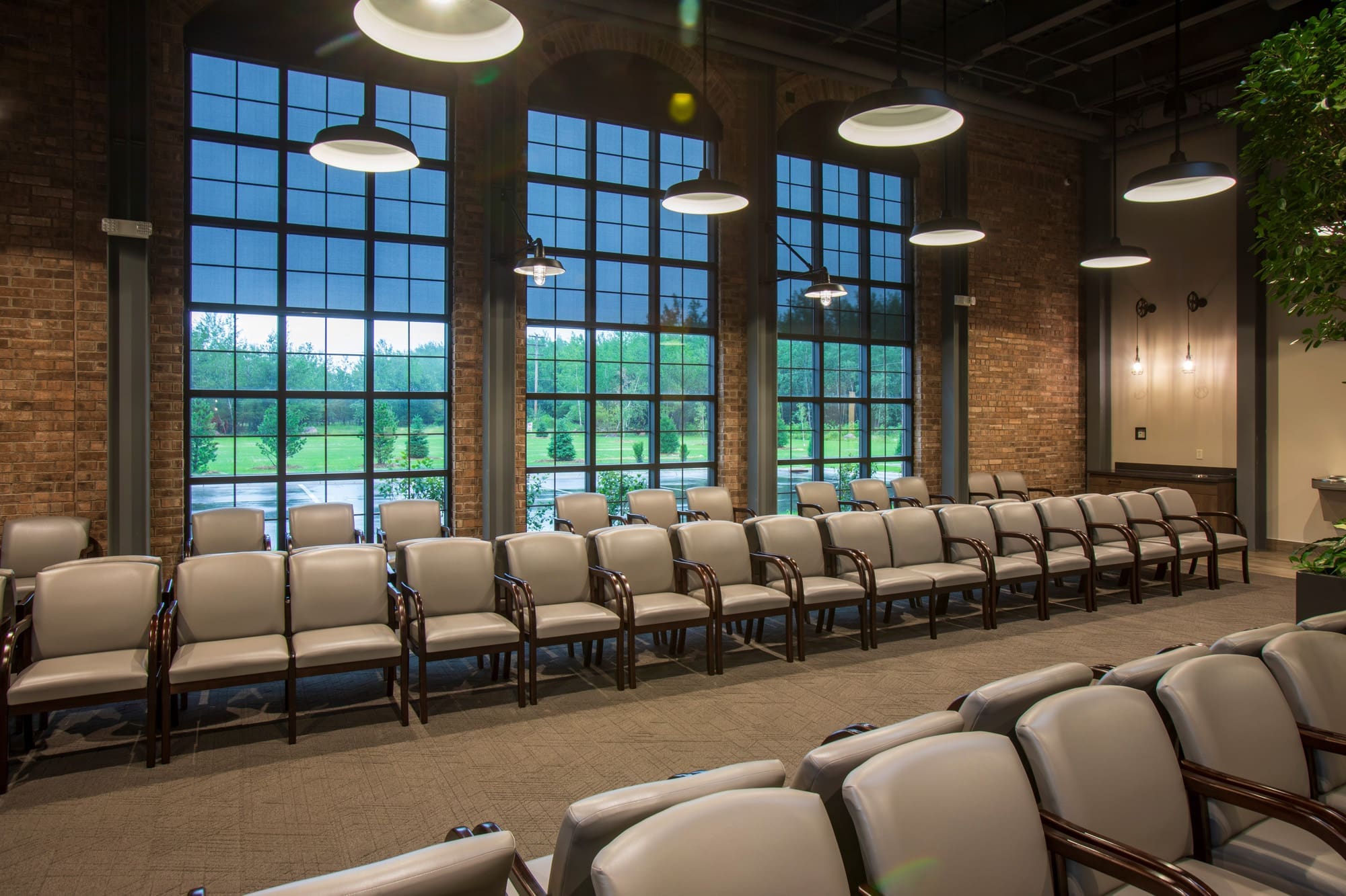 Spacious commercial meeting room with rows of beige chairs, brick walls, industrial windows, and modern lighting, ideal for events.