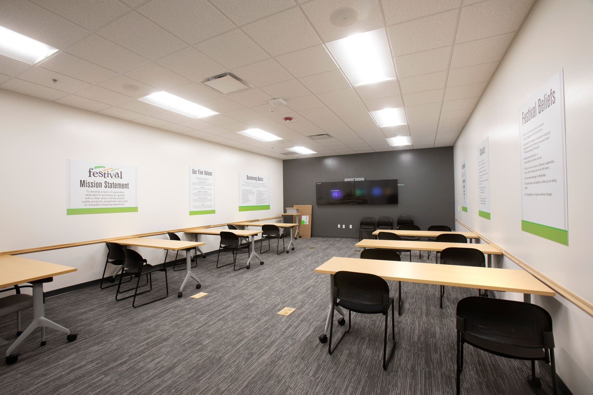 Commercial classroom space featuring light wood tables, black chairs, bright ceiling lights, and a large display monitor.